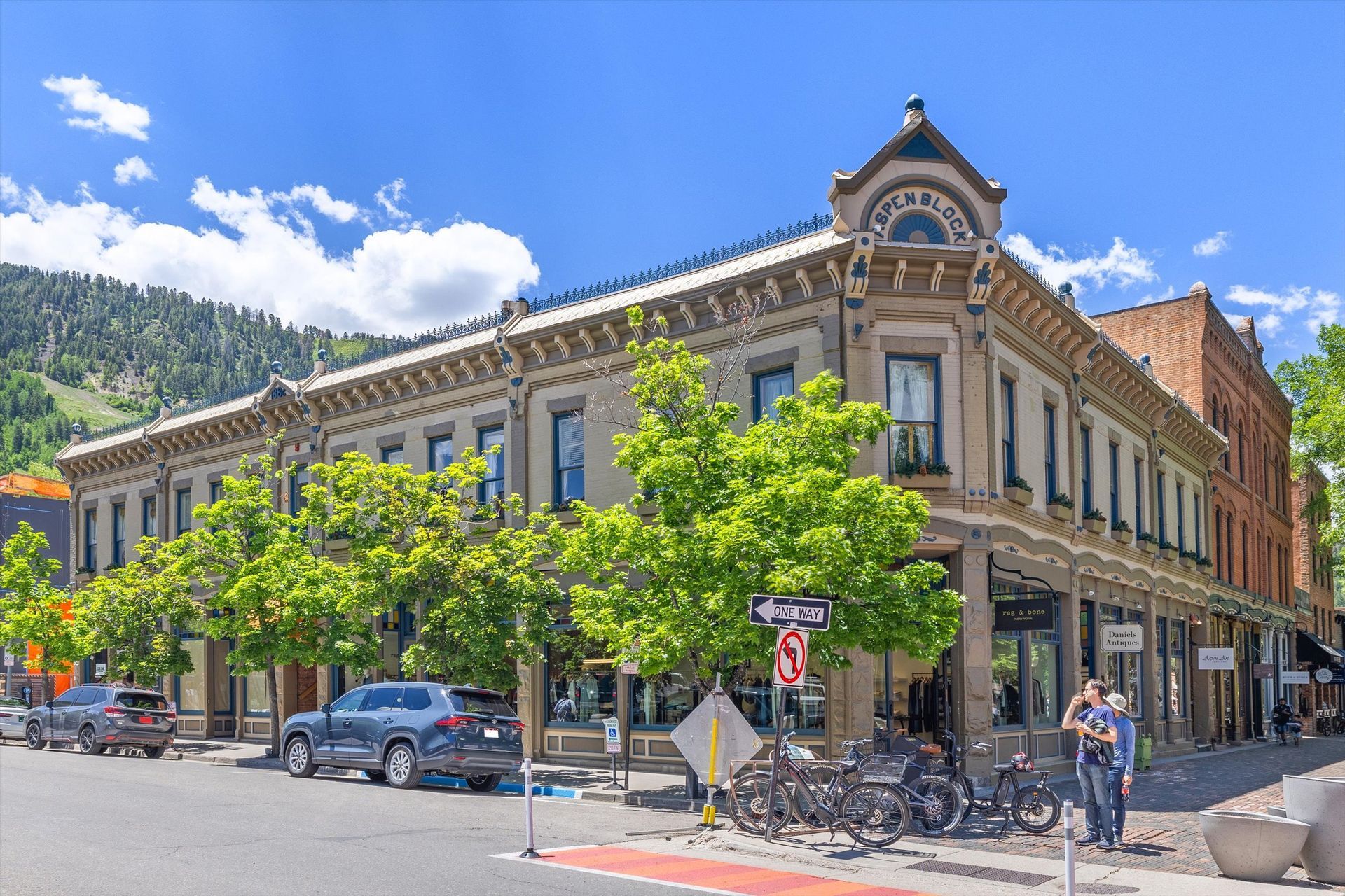 Historic building on a street corner, with parked cars, bicycles, and pedestrians. Green trees, blue sky.