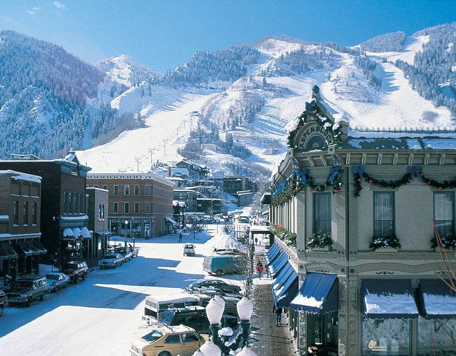 Snow-covered street in a mountain town with buildings and a ski slope in the background.