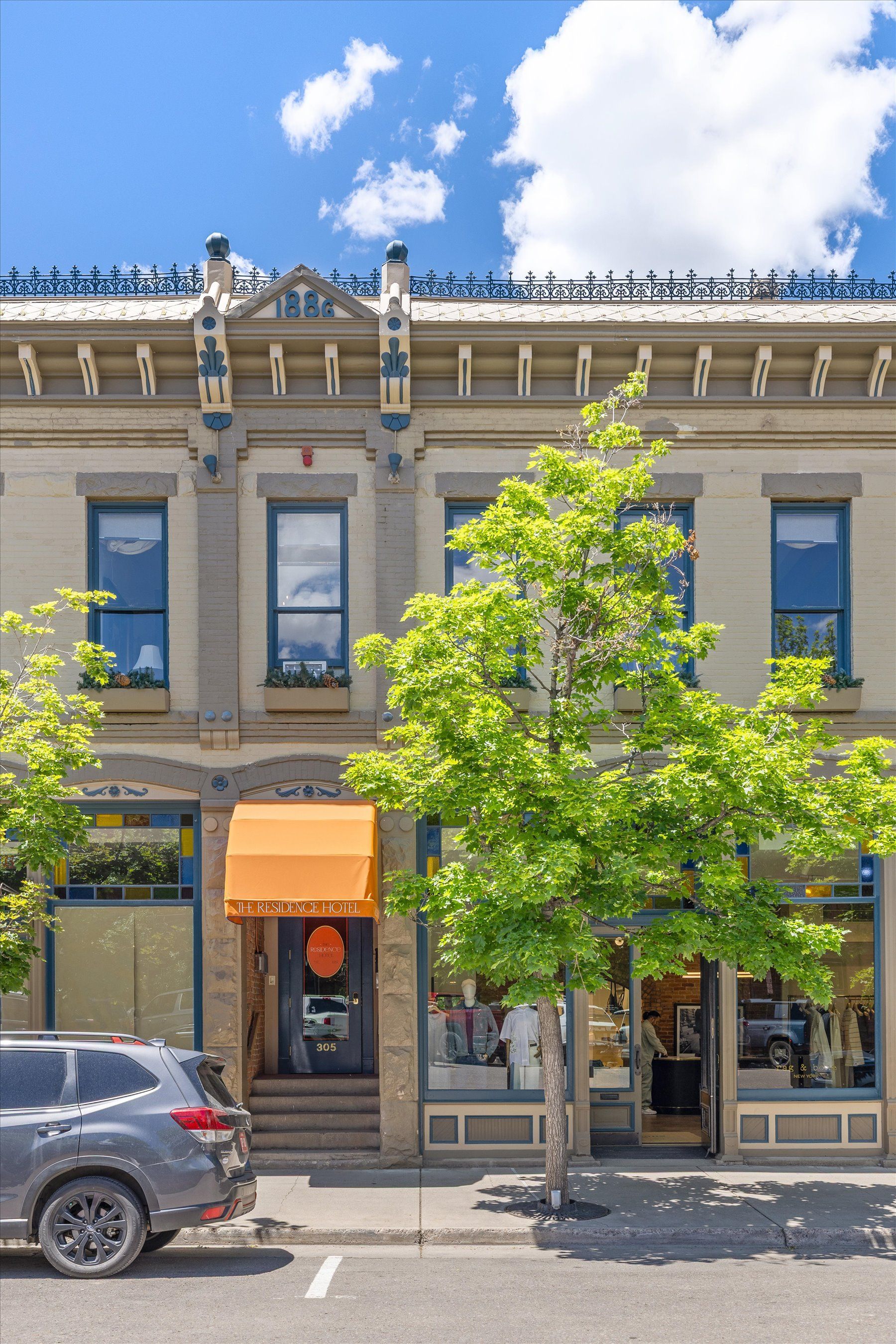 Two-story brick building with businesses on the ground floor, orange awning, and a tree in front. Blue sky overhead.
