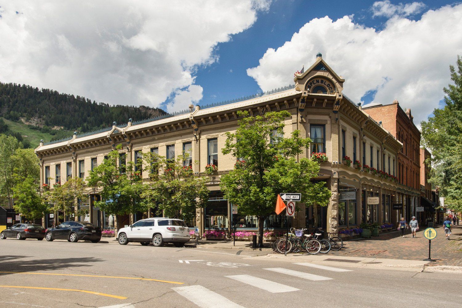 Historic two-story building in Aspen, Colorado. Trees line the sidewalk, cars parked nearby. Sunny day, blue sky.