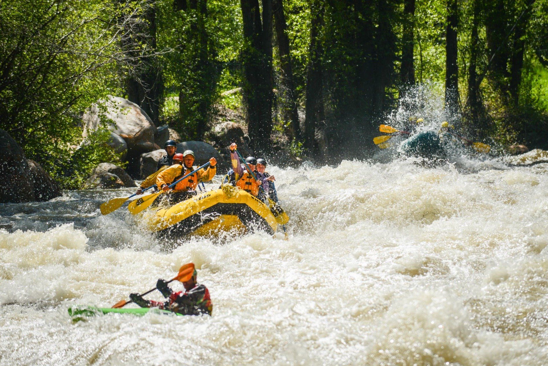 Rafting in whitewater rapids; yellow raft with multiple people, one kayaking in choppy water.