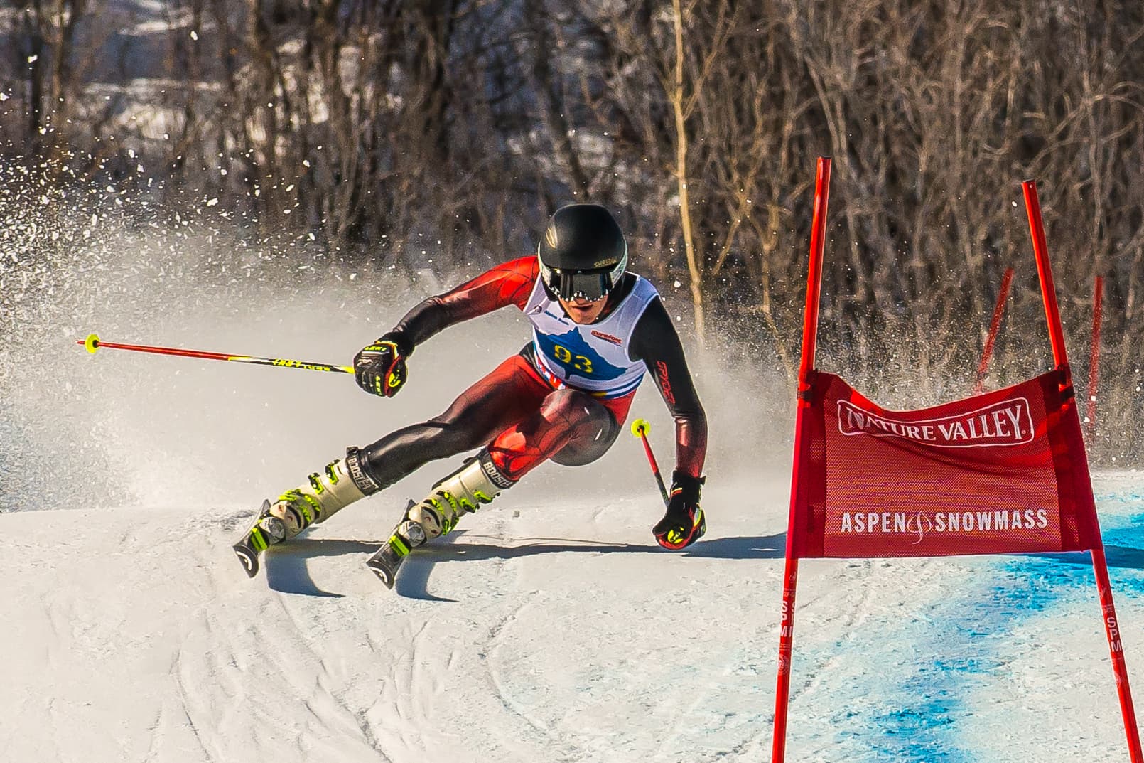 Skier in black and red racing suit, skiing downhill past red course marker. Snow flies.