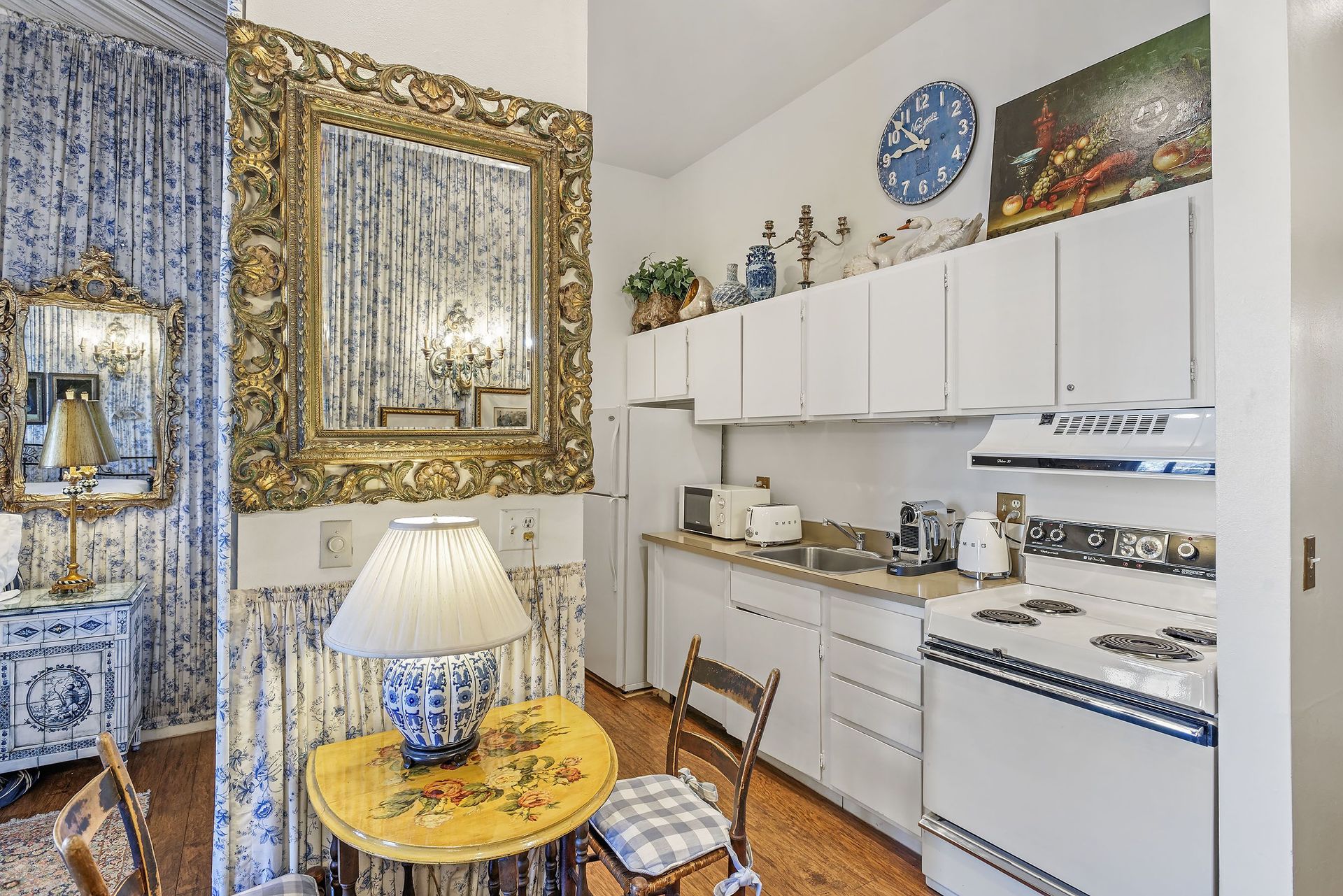 Small, ornate kitchen with white cabinets, appliances, and walls, decorated with an antique mirror and table.