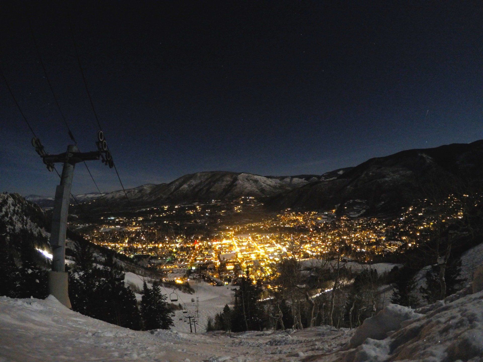 Nighttime view of a brightly lit town nestled in a snowy mountain valley, with a ski lift tower in the foreground.