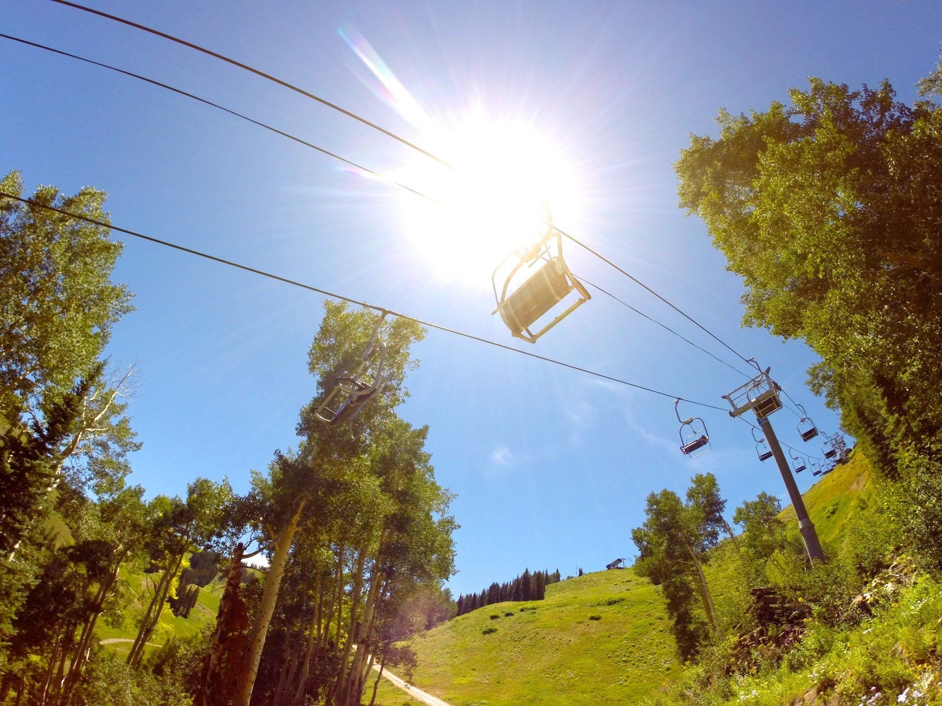 Ski lift ascending a sunny hillside, with a bright sun overhead. Green trees and blue sky.