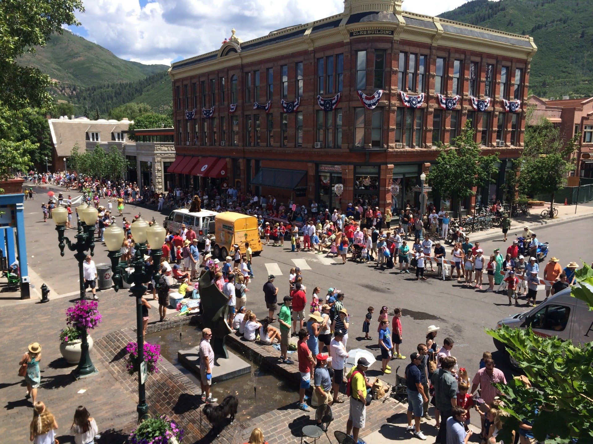 Street parade in front of a brick building; crowd watches, buildings, green mountains in the background.