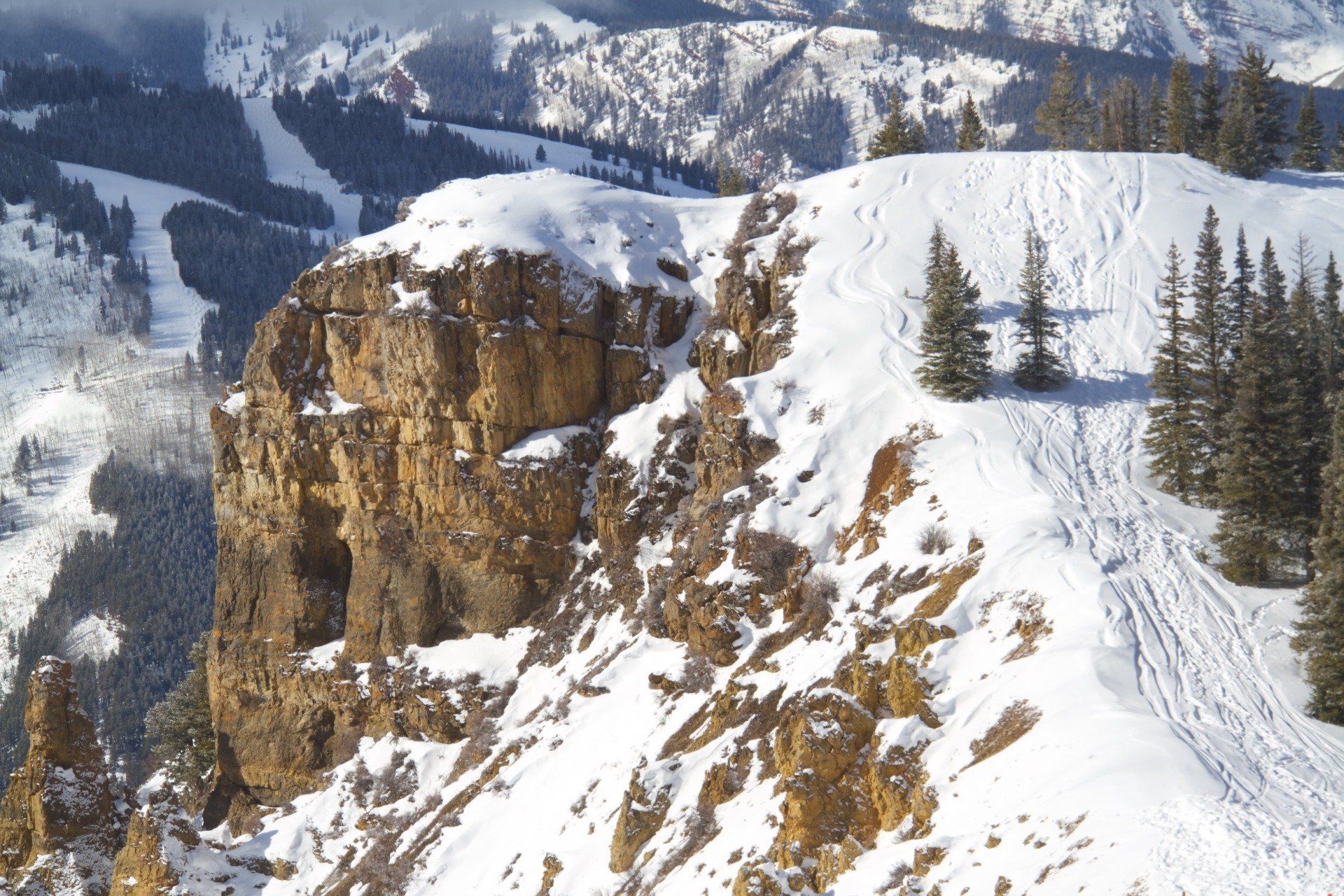 Snow-covered mountain ridge with a rocky cliff face, trees, and ski runs visible in the background.