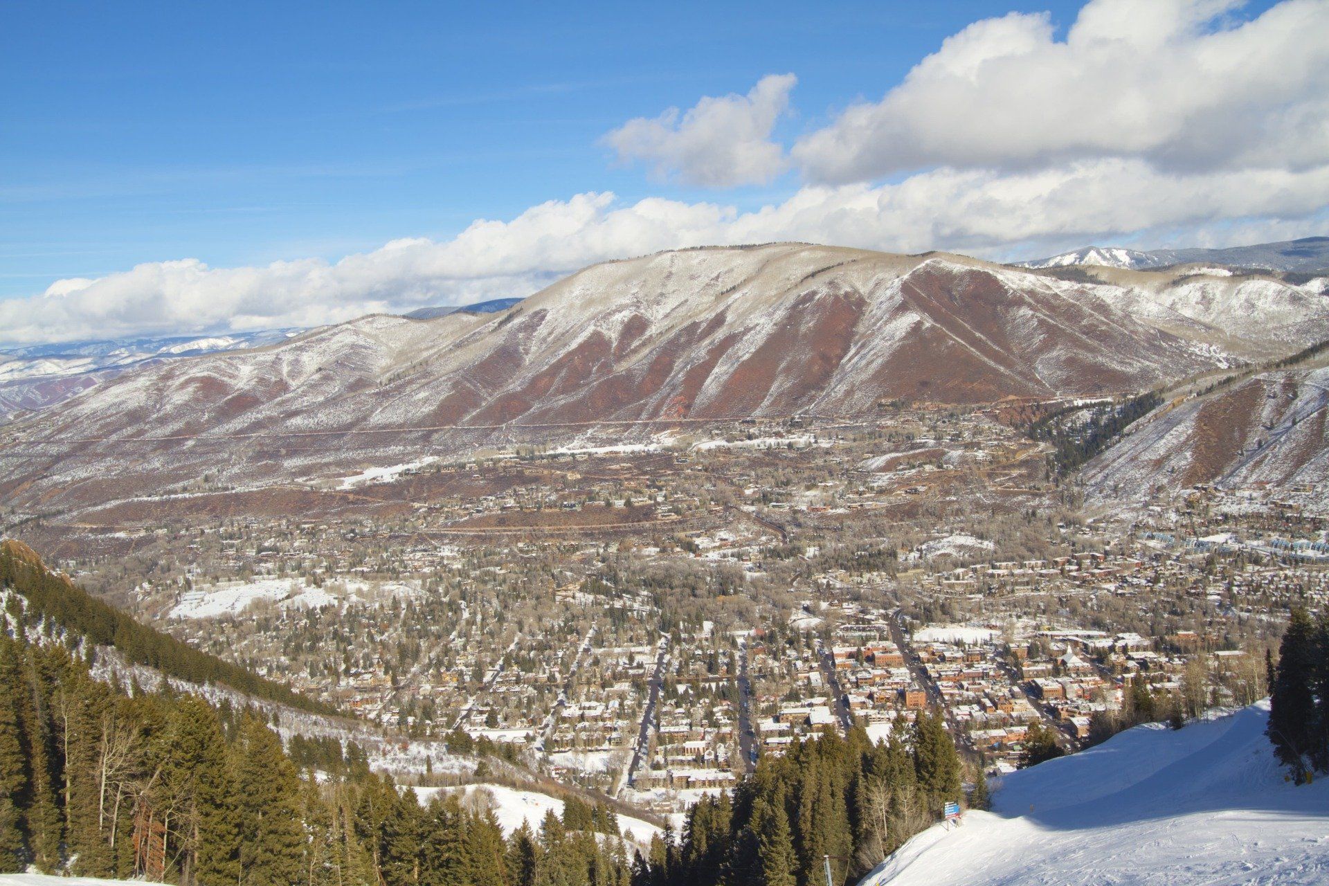 Snowy mountain view overlooking a town; brown mountain, green trees, blue sky.