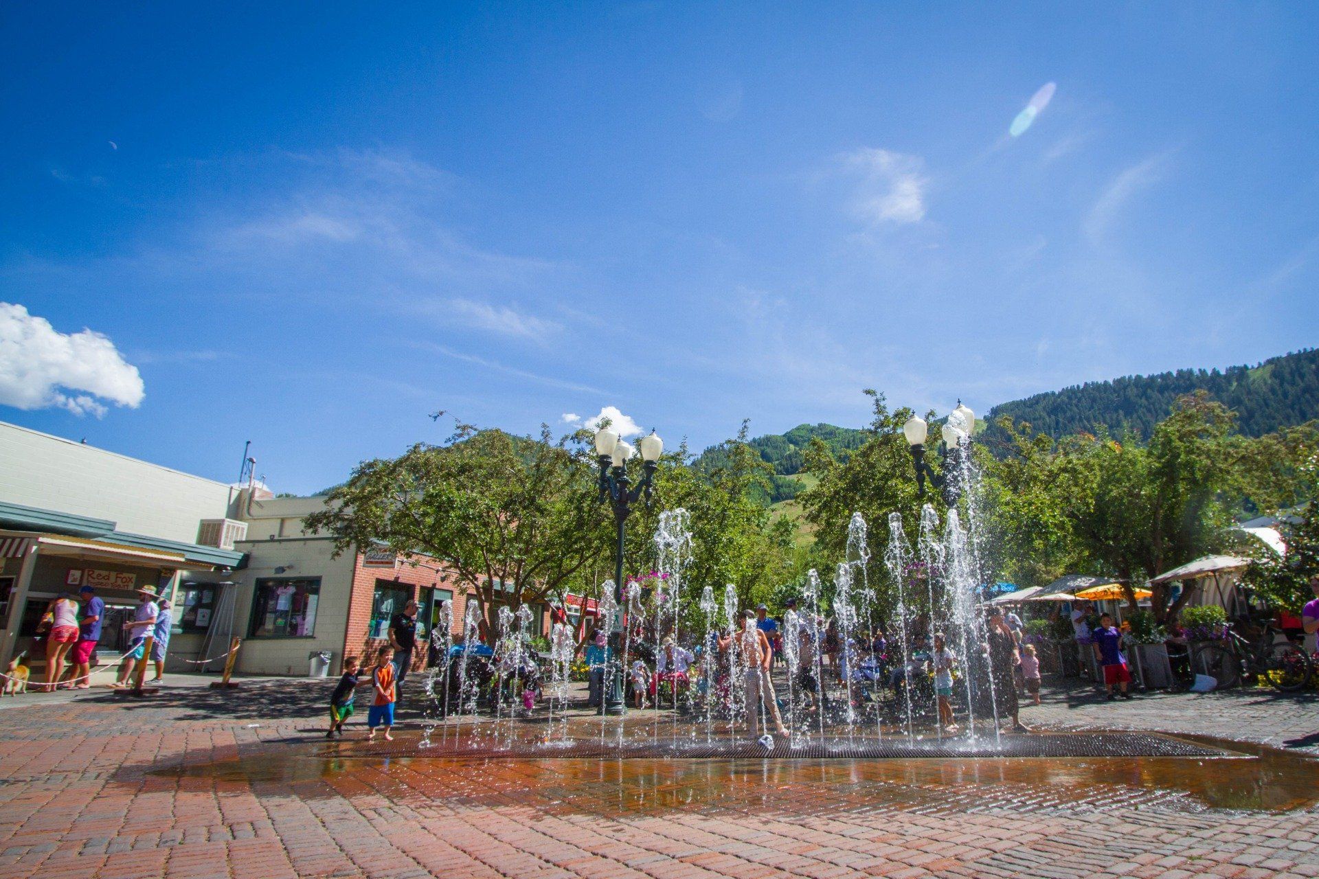 Fountain spraying water in a town square. People gather around shops and trees on a sunny day.