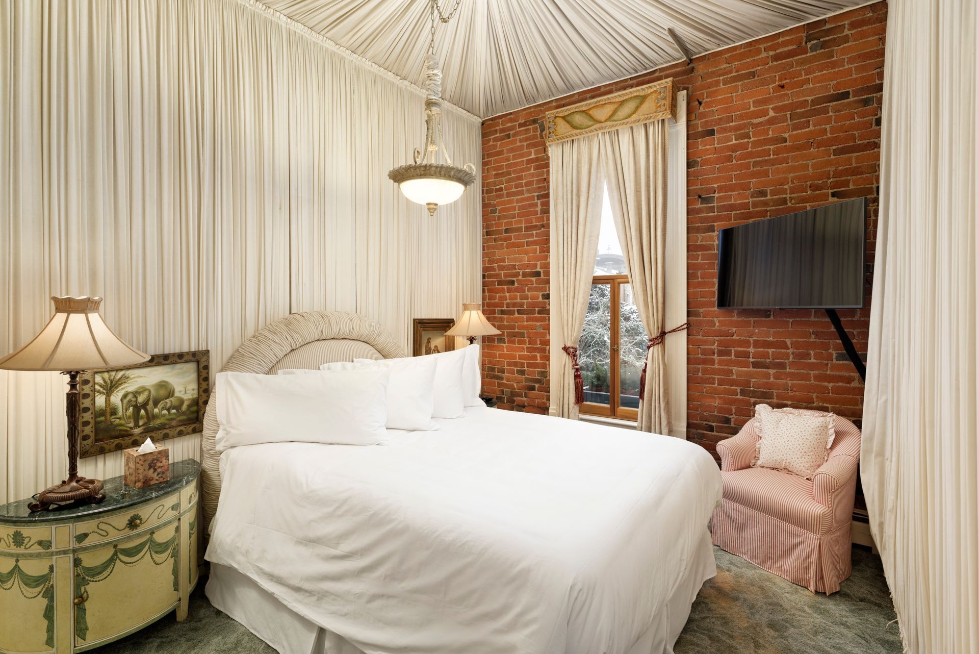 Bedroom with a white bed, exposed brick wall, draped curtains, and a mounted TV.