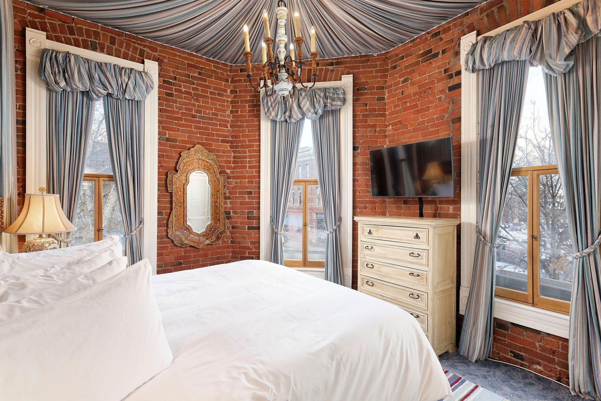 Bedroom with exposed brick walls, white bedding, and ornate gold mirror. Blue curtains frame windows.