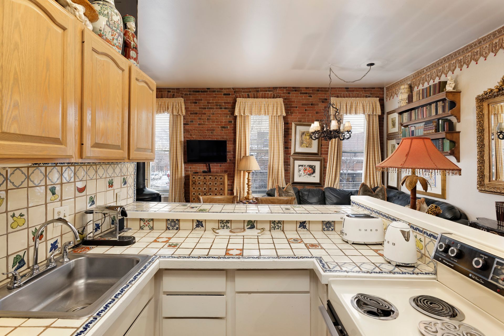 Kitchen with light wood cabinets, tiled backsplash and countertops, brick wall, and small windows.