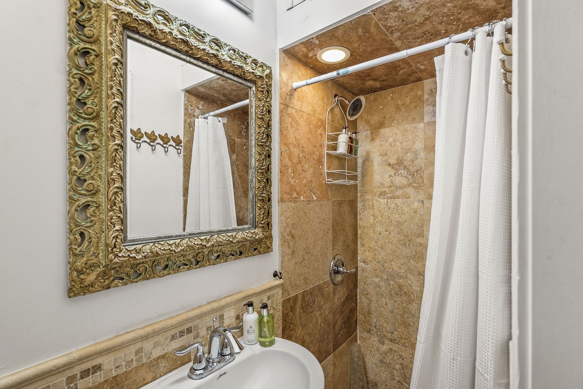 Bathroom with a shower, sink, and large ornate mirror. Beige tile, white shower curtain, and a gold-framed mirror.
