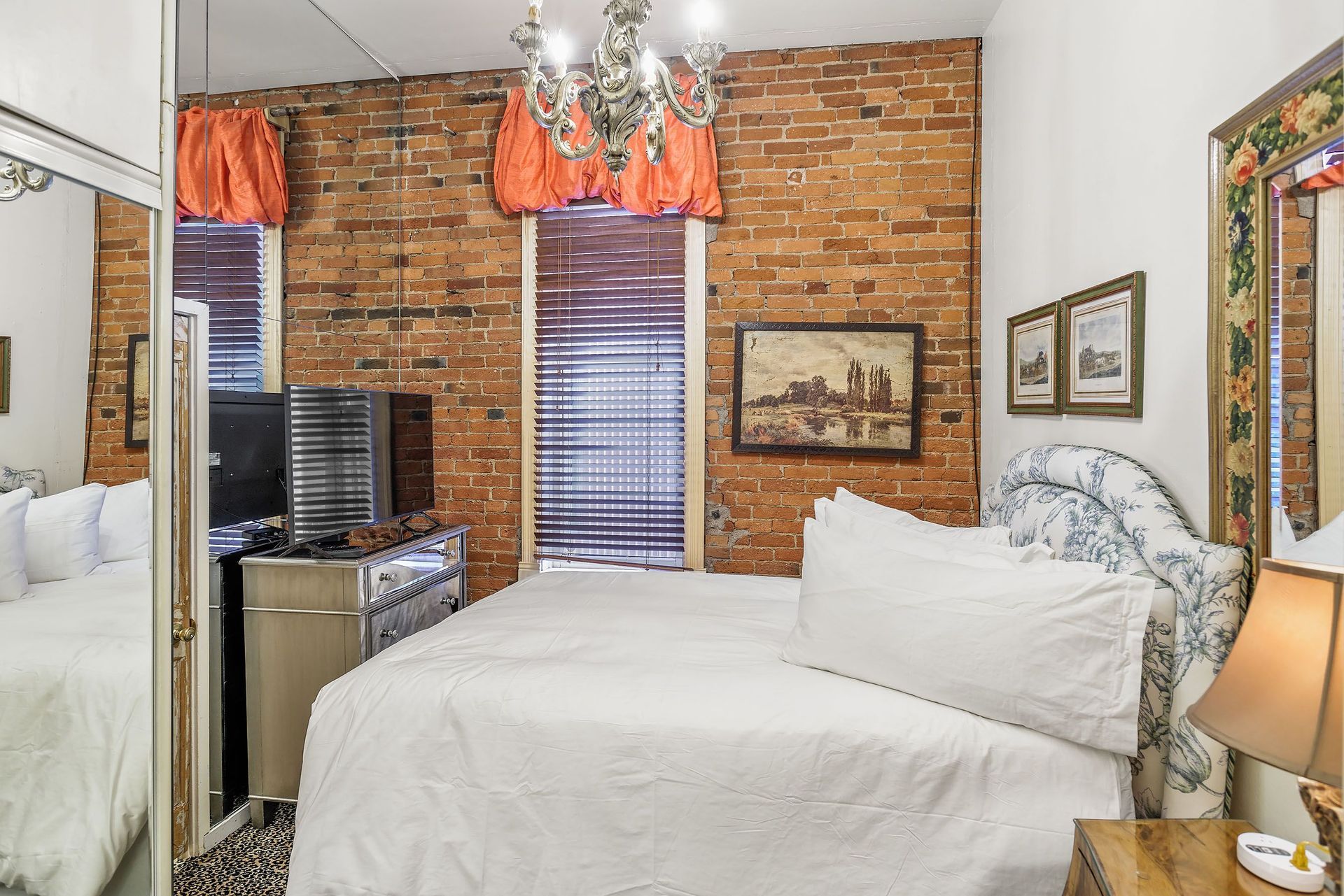 Bedroom with exposed brick wall, bed, dresser, window with curtains, and chandelier.