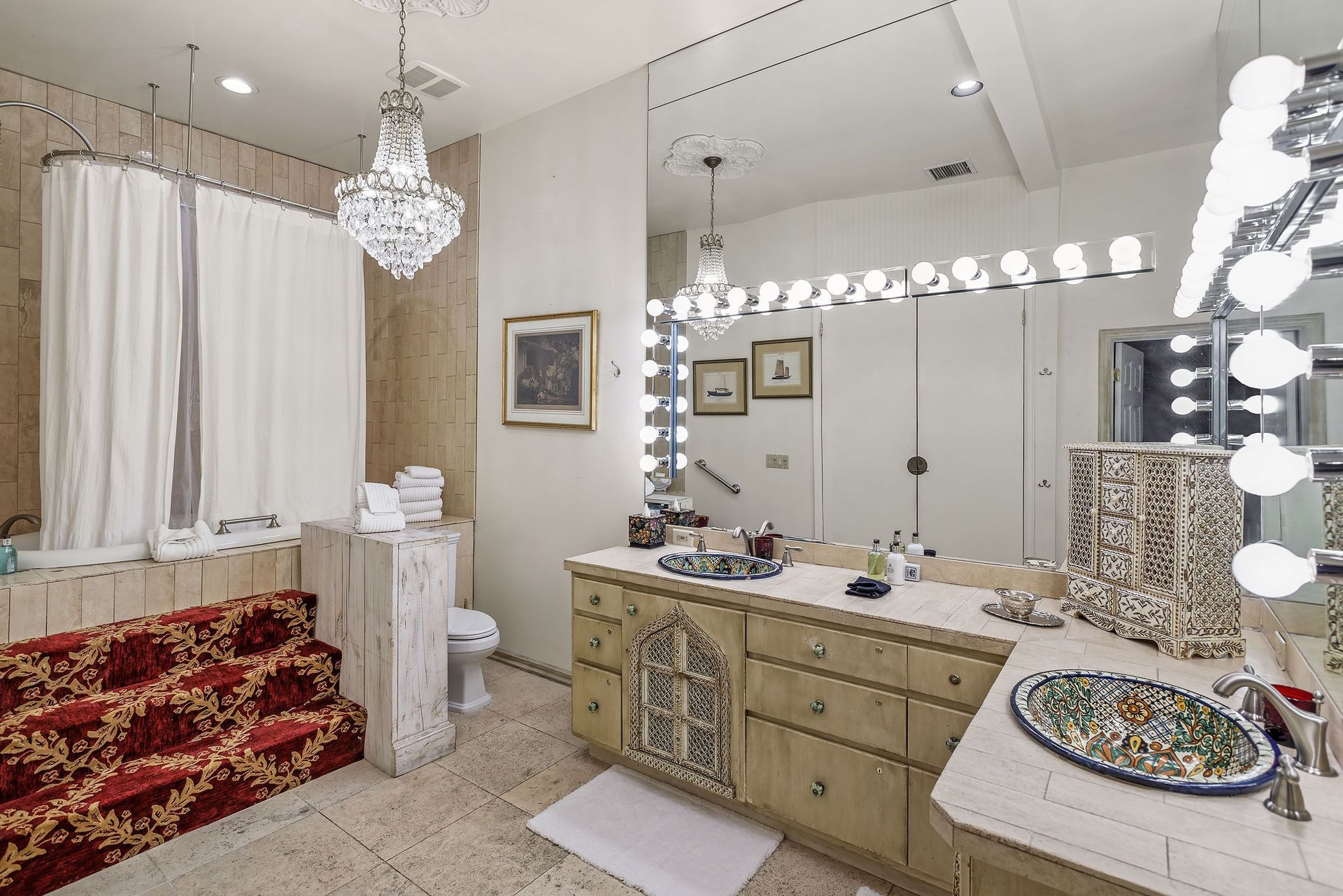 Bathroom with ornate light fixtures, beige and red accents, large mirror, and decorative sink.