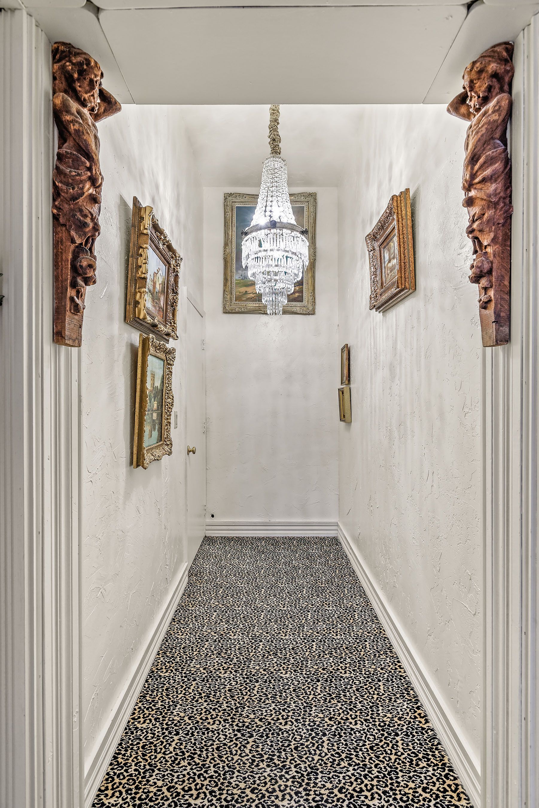Narrow hallway with ornate molding, artwork, a chandelier, and patterned carpet.