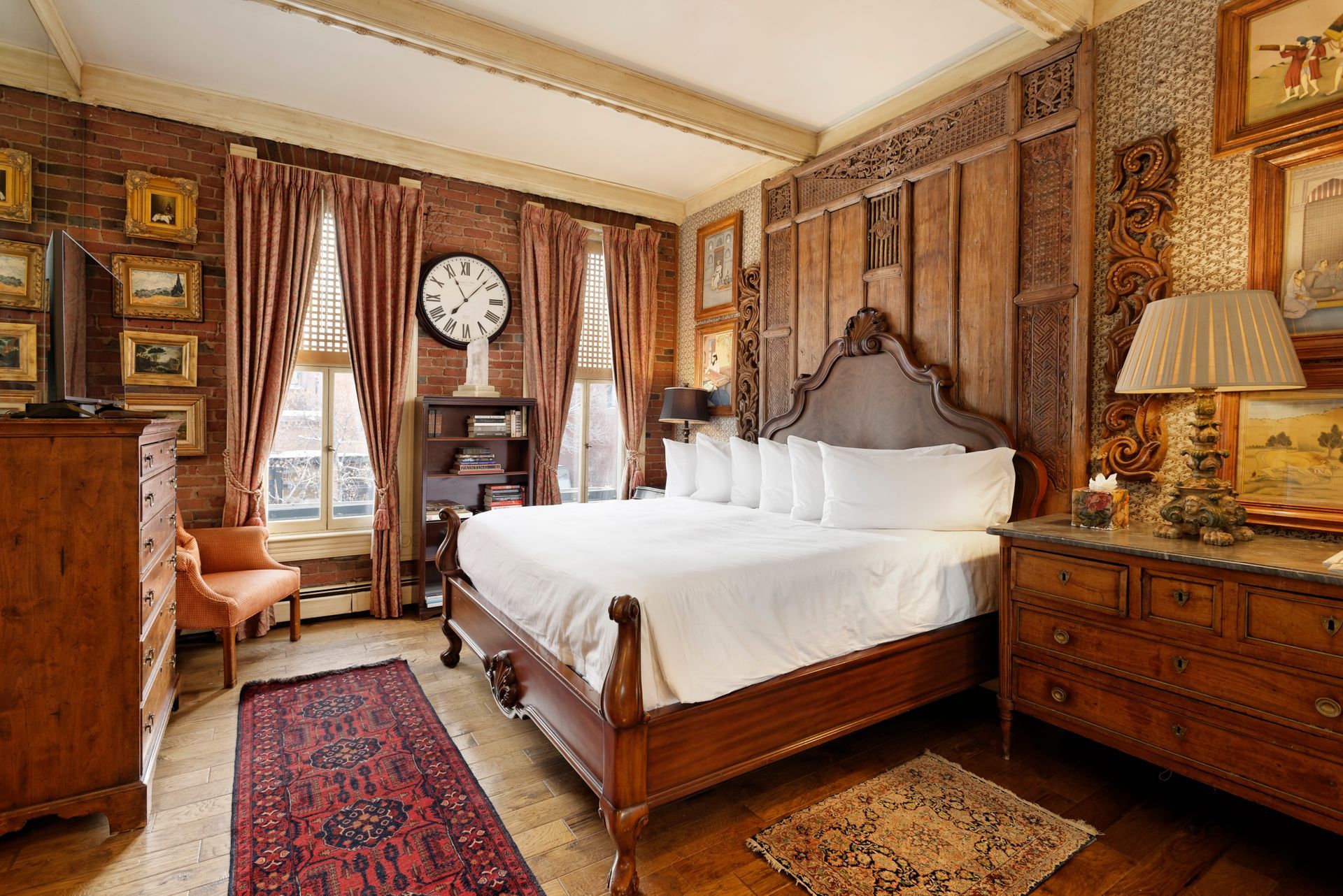 Bedroom with exposed brick walls, antique furniture, ornate headboard, framed art, and a large clock.