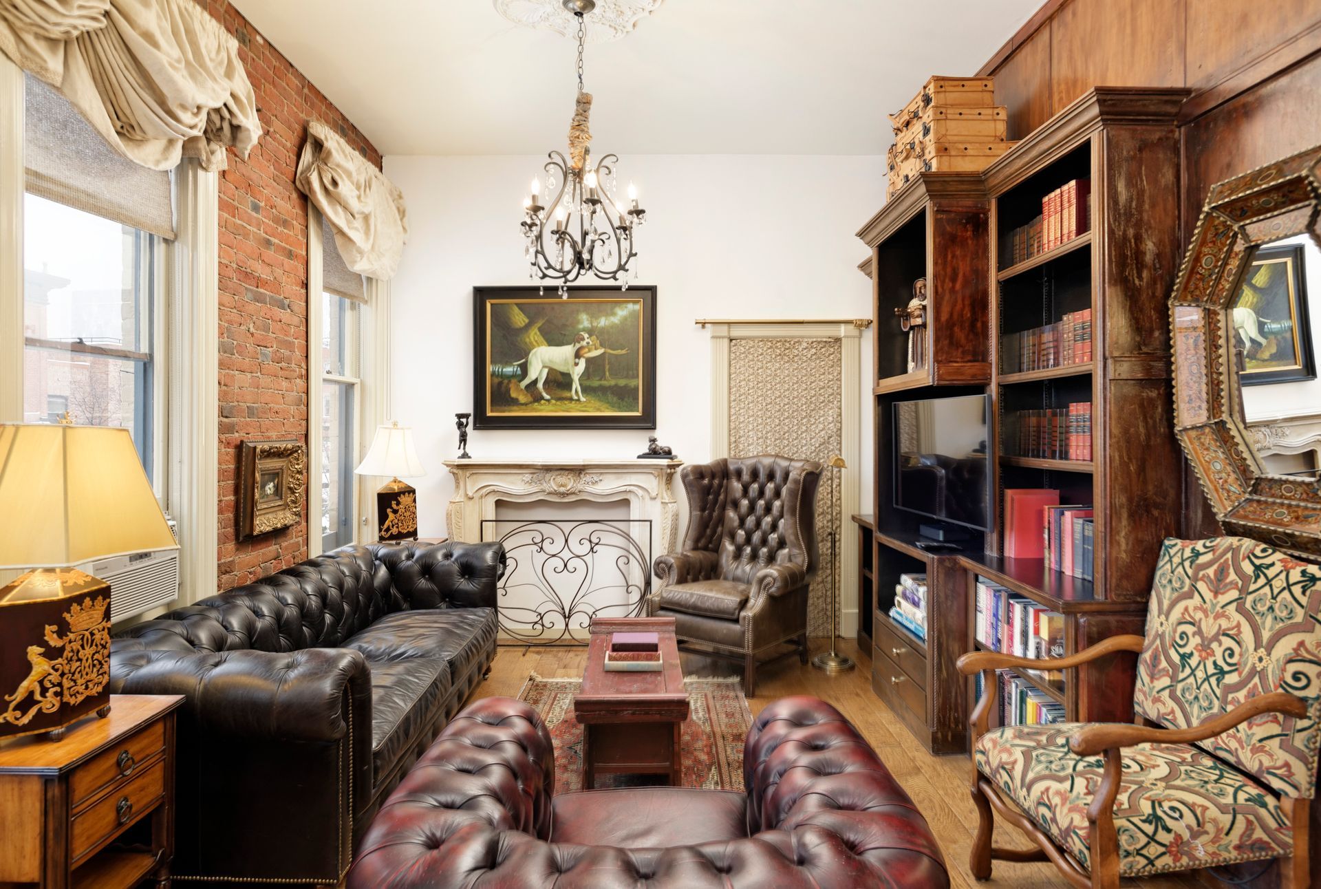 Cozy living room with dark leather Chesterfield sofas, brick wall, fireplace, and bookshelves.