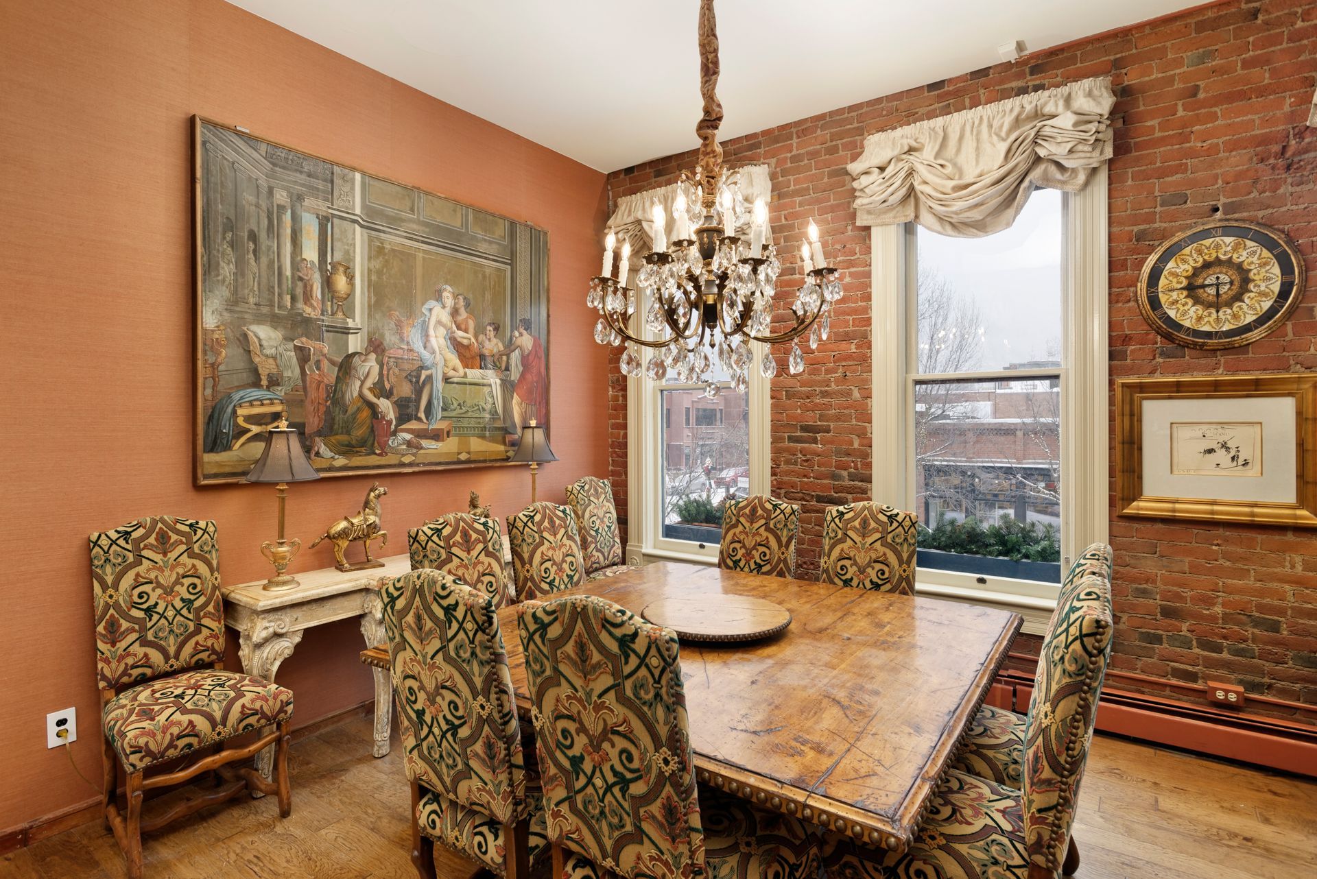 Dining room with brick wall, chandelier, large artwork, and ornate dining table with chairs.