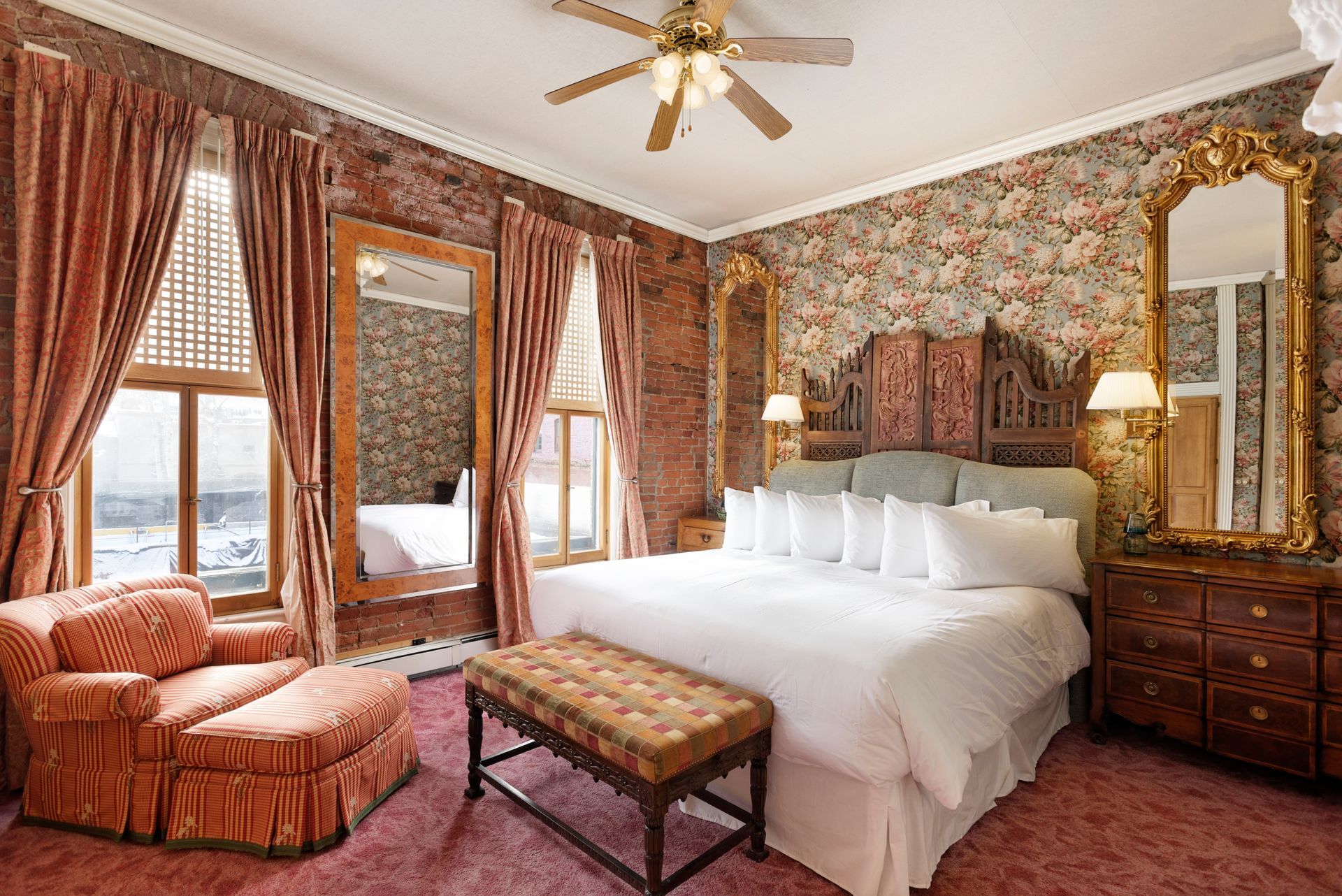 Bedroom with floral wallpaper, brick walls, and a large bed with a bench. An armchair sits near a window.