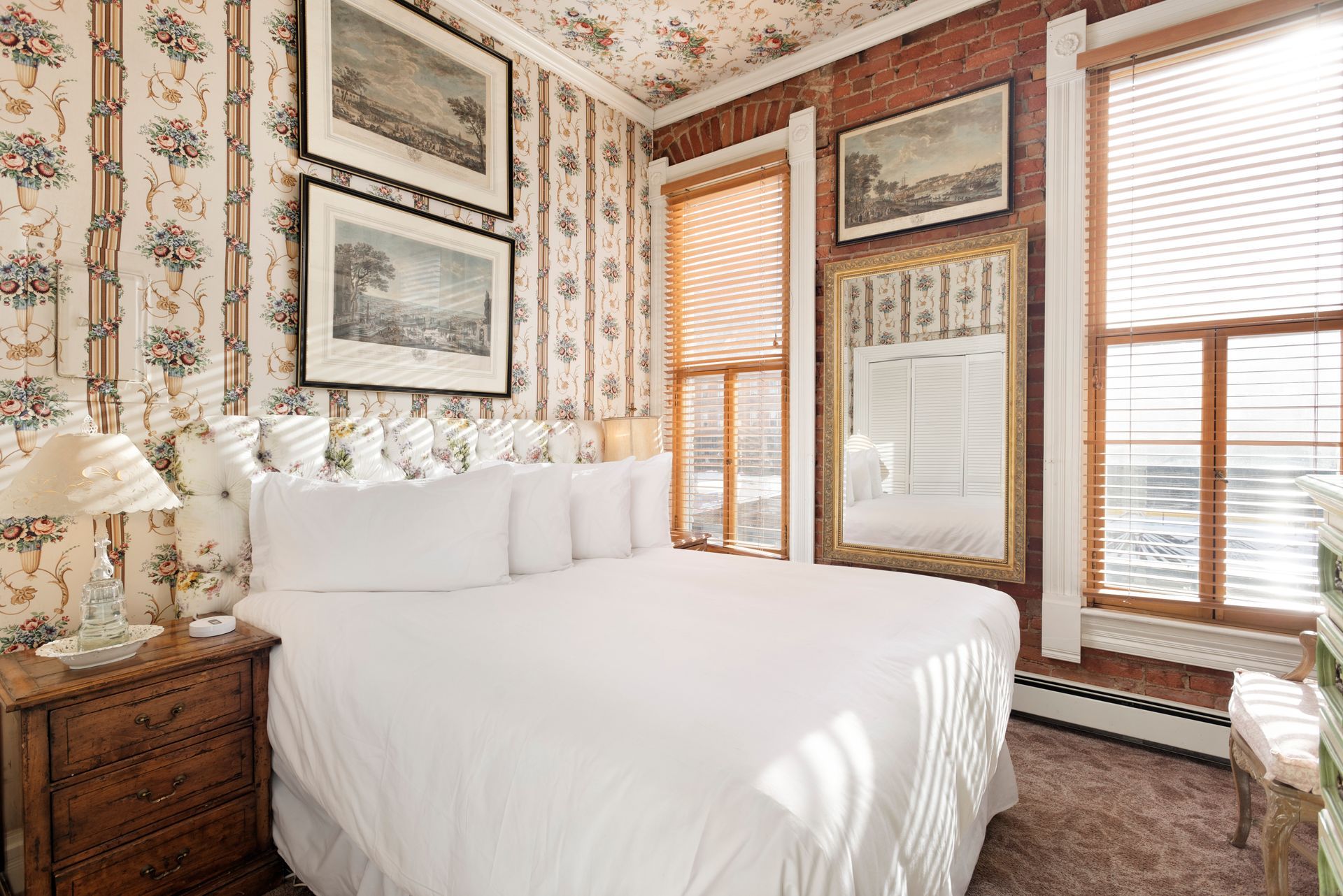 Bedroom with floral wallpaper, bed, nightstand, framed art, and window with blinds.