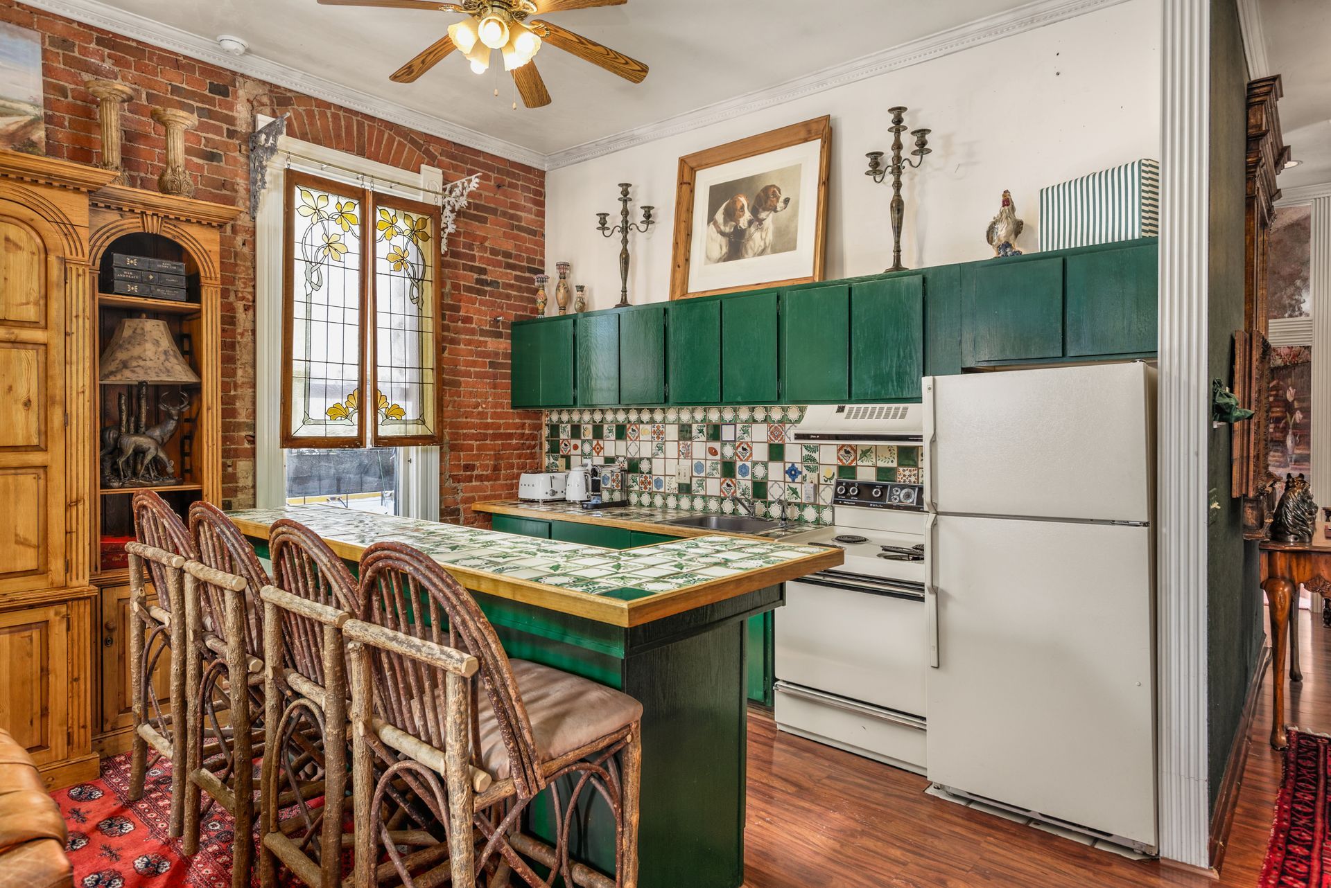 Kitchen with brick wall, green cabinets, tiled countertop, and wicker bar stools.