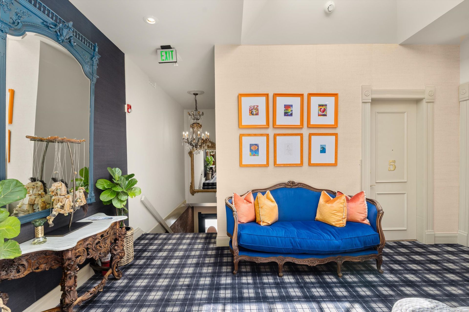 Blue couch with orange pillows, framed art, antique table with mirror, plaid carpet, and a chandelier.