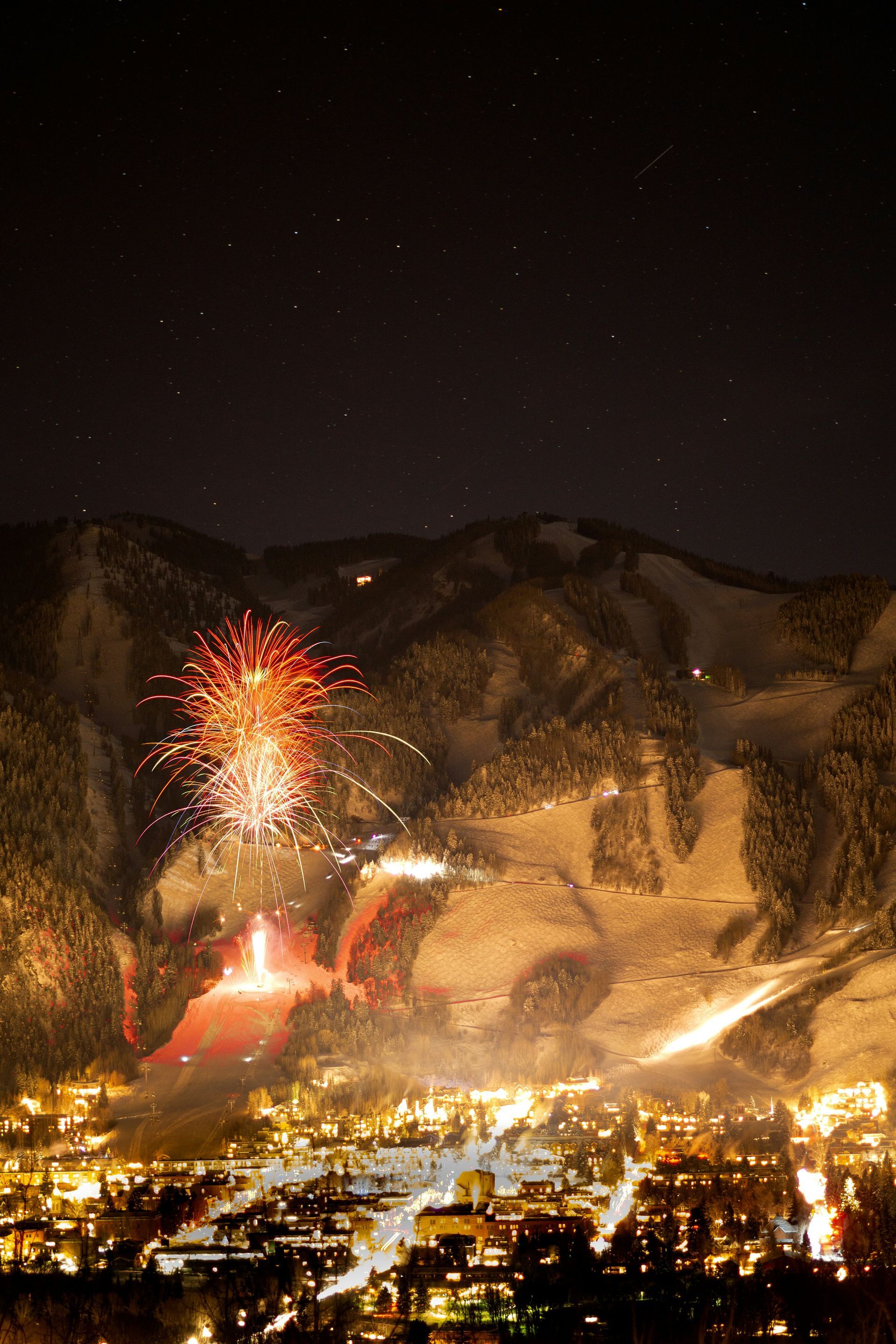 Fireworks exploding over a snow-covered mountain, illuminating a town at night.