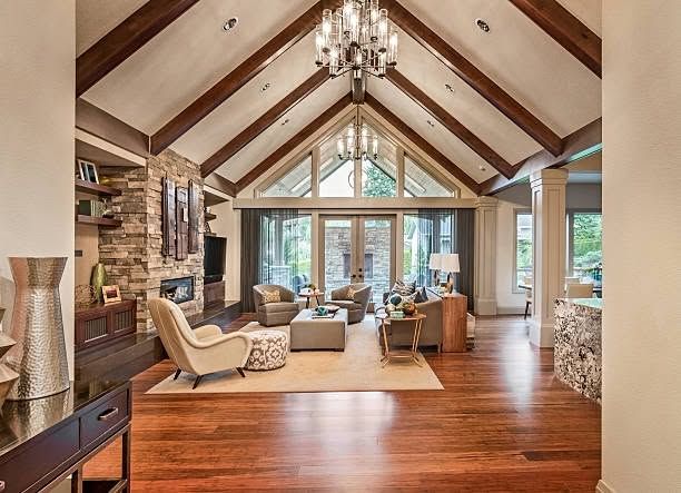 Spacious living room with vaulted wood ceiling, stone fireplace, and large windows overlooking trees.