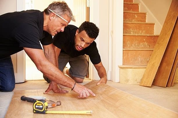 Two people measuring wood flooring in a room, with tools on the floor and stairs in the background.