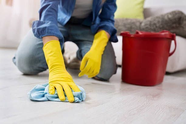 Person in yellow gloves cleaning a light wood floor with a cloth beside a red bucket.