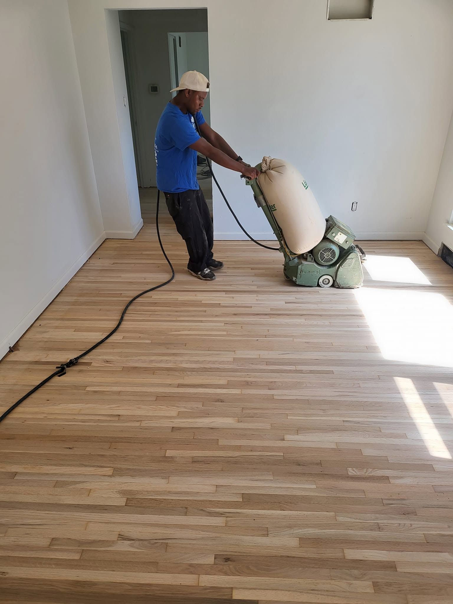 Worker sanding a hardwood floor with a floor sander in a nearly empty room