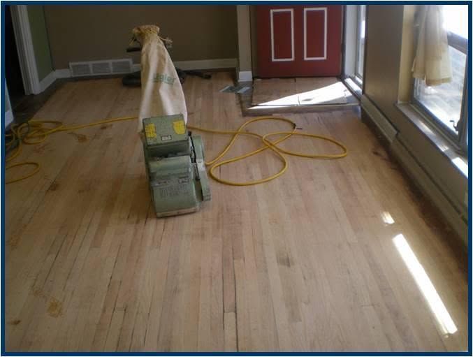 Worker sanding a wooden floor in a room with yellow extension cords and a red door.