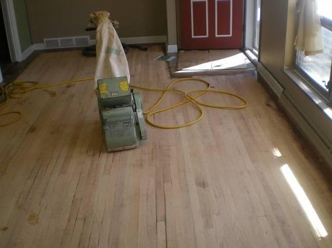 Wood flooring being sanded with a floor sander and yellow extension cord in a room with a red door.