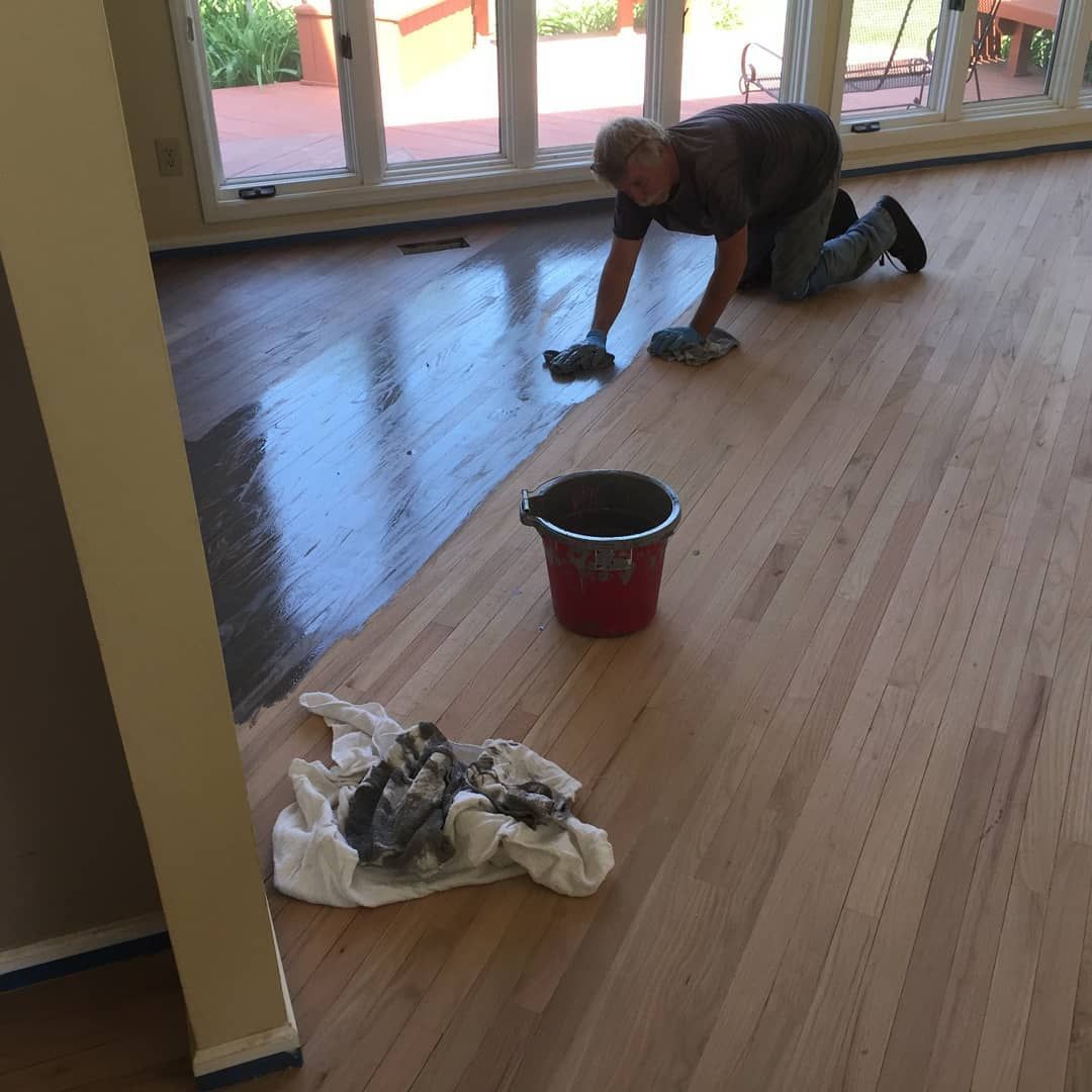 Worker refinishing a hardwood floor with a bucket and rag in a room with large windows
