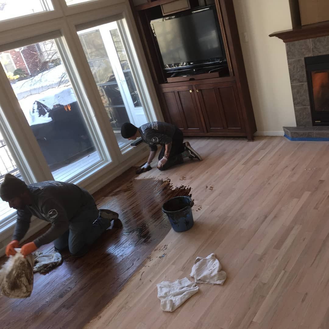 Two people cleaning a hardwood living room floor near sliding glass doors and a TV.