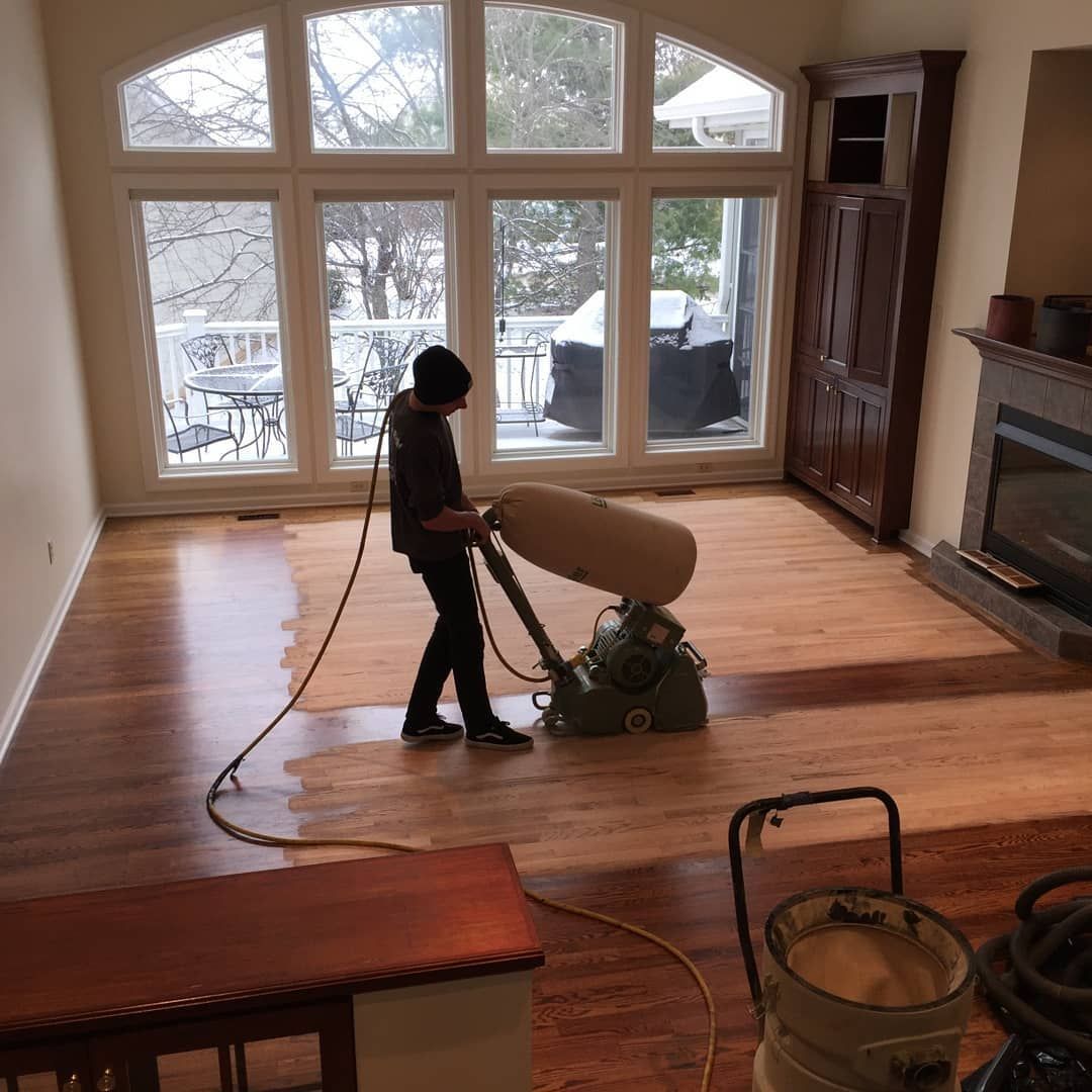 Person sanding a hardwood floor in a living room with large windows and a fireplace.