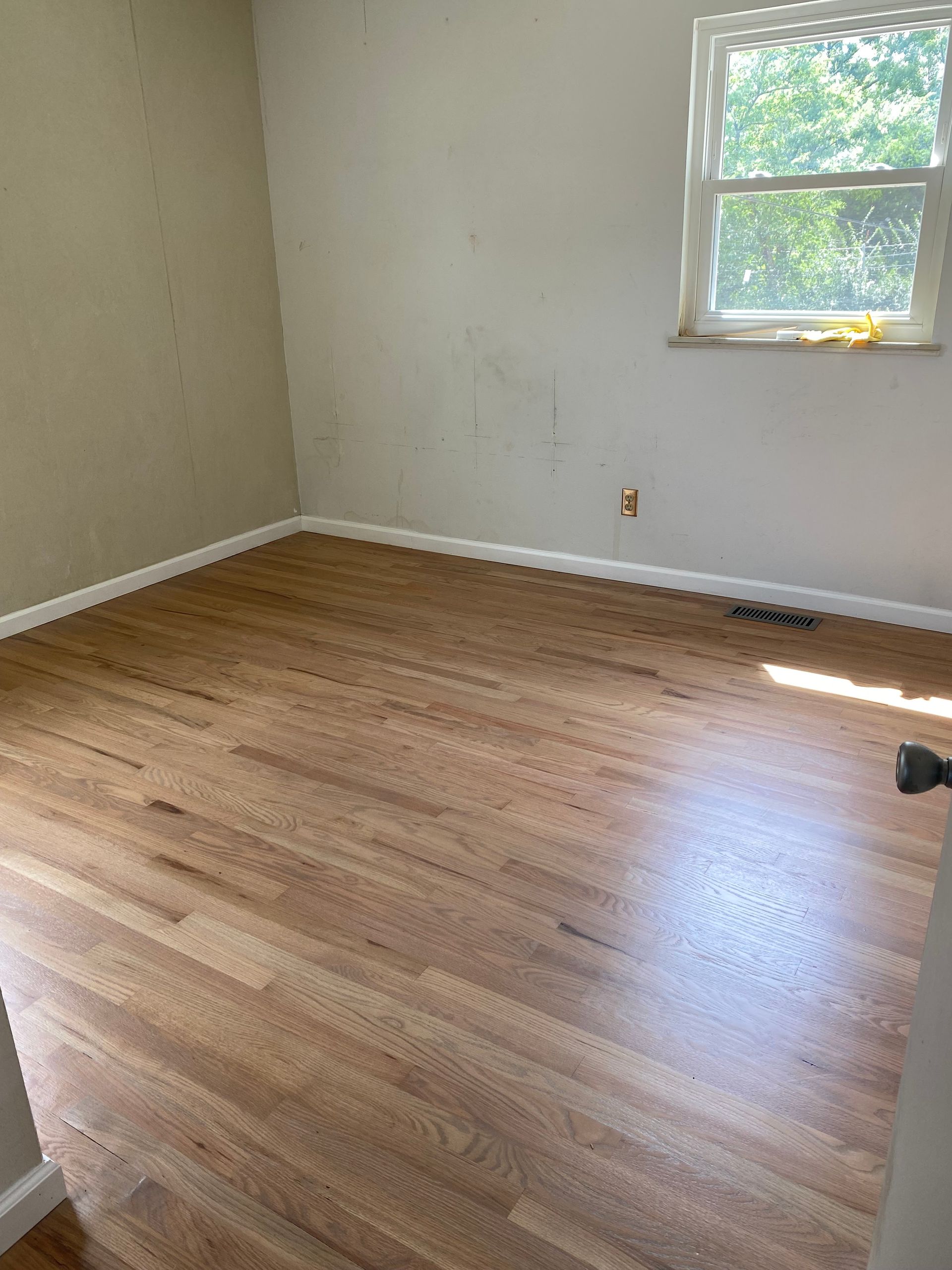 Empty room with light wood floors, beige walls, and a sunlit window.