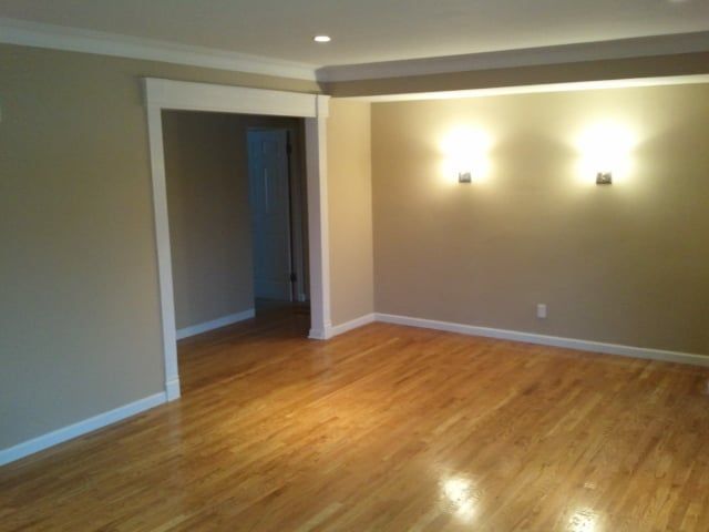 Empty living room with hardwood floors, beige walls, and two wall sconces lit near an open doorway.