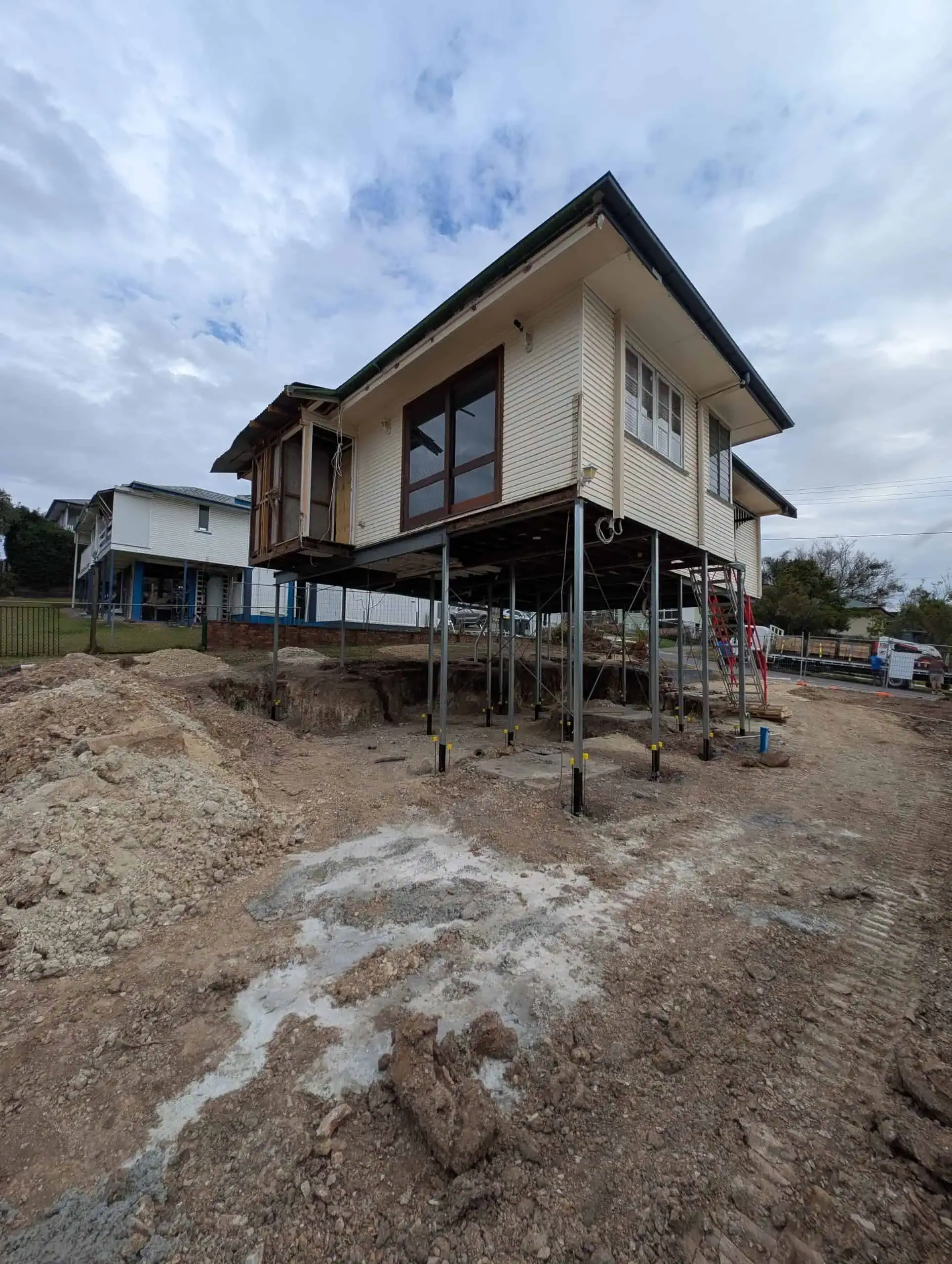 Raised house on stilts over rough, muddy ground under a cloudy sky, with another house in the background