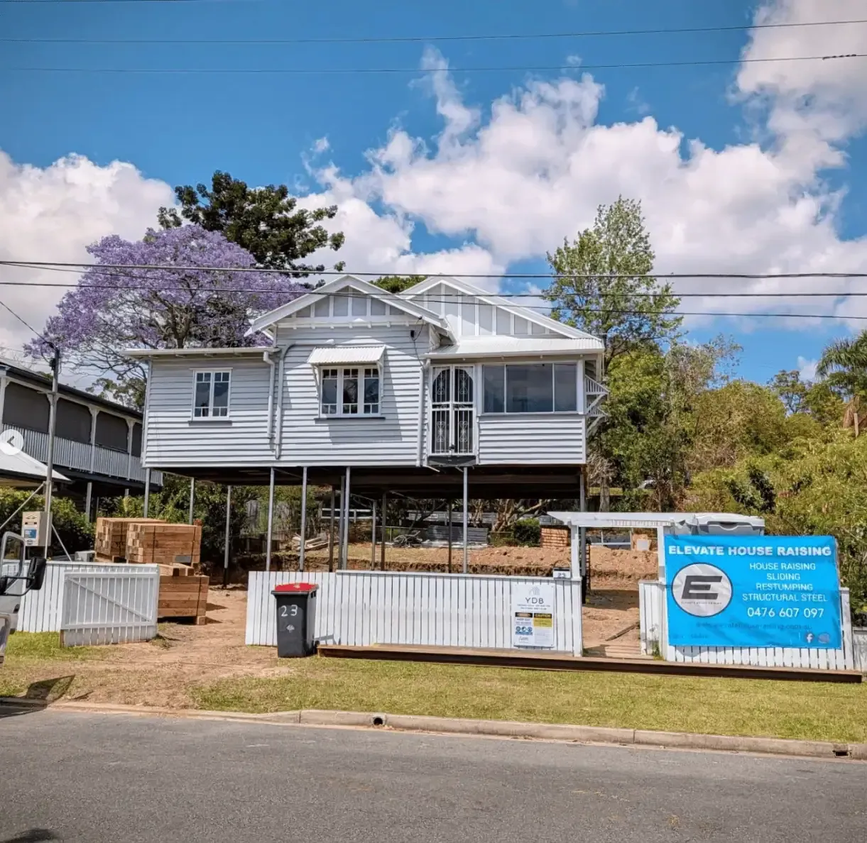 Raised white house under construction with a front fence, trees, and a blue sign by the road