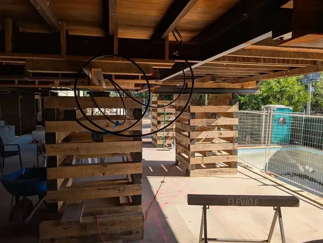 Wooden deck under construction with exposed beams, stacked lumber, and a wire fence in bright daylight