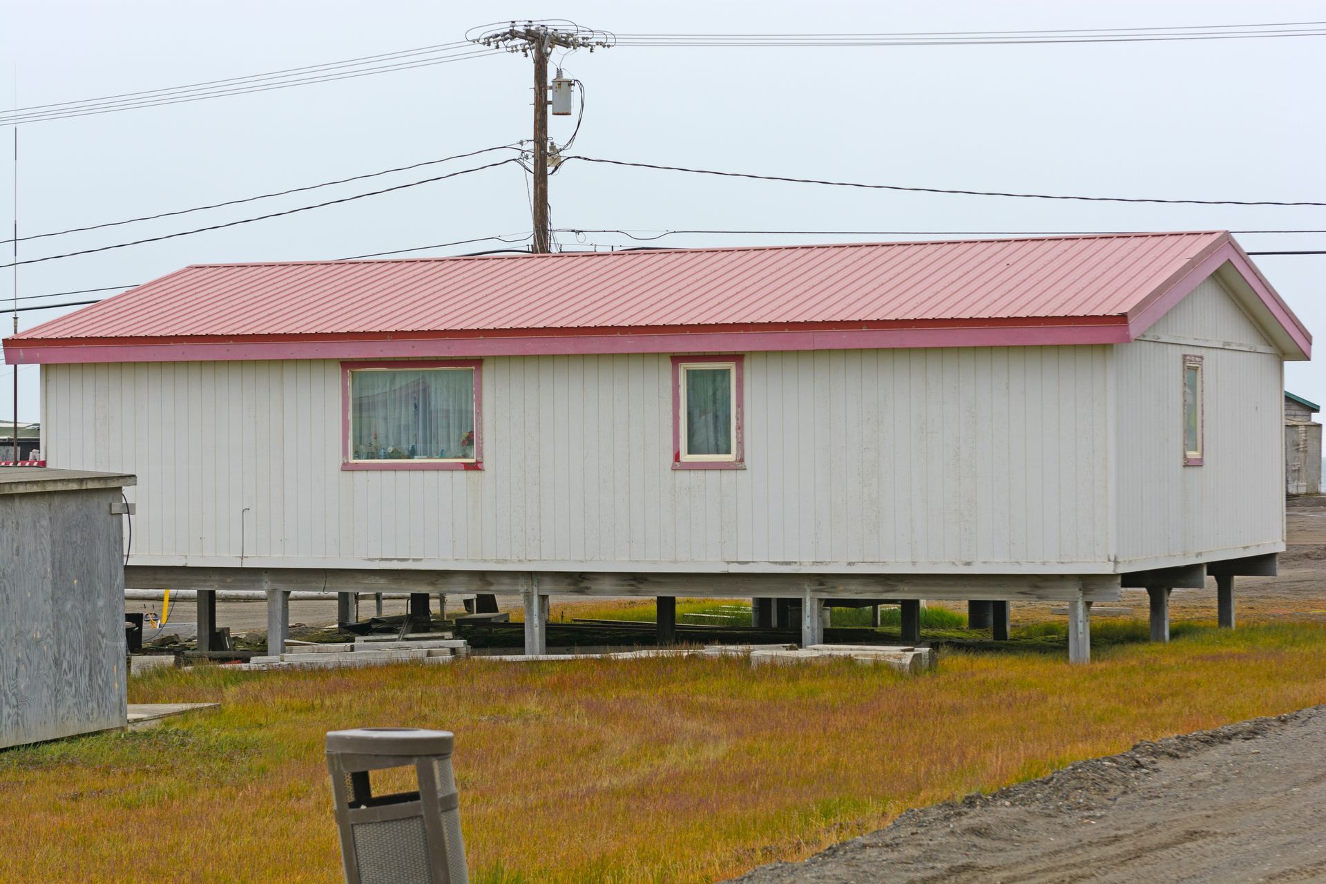 Small white elevated house with red roof on a cloudy day, beside a road and utility pole.
