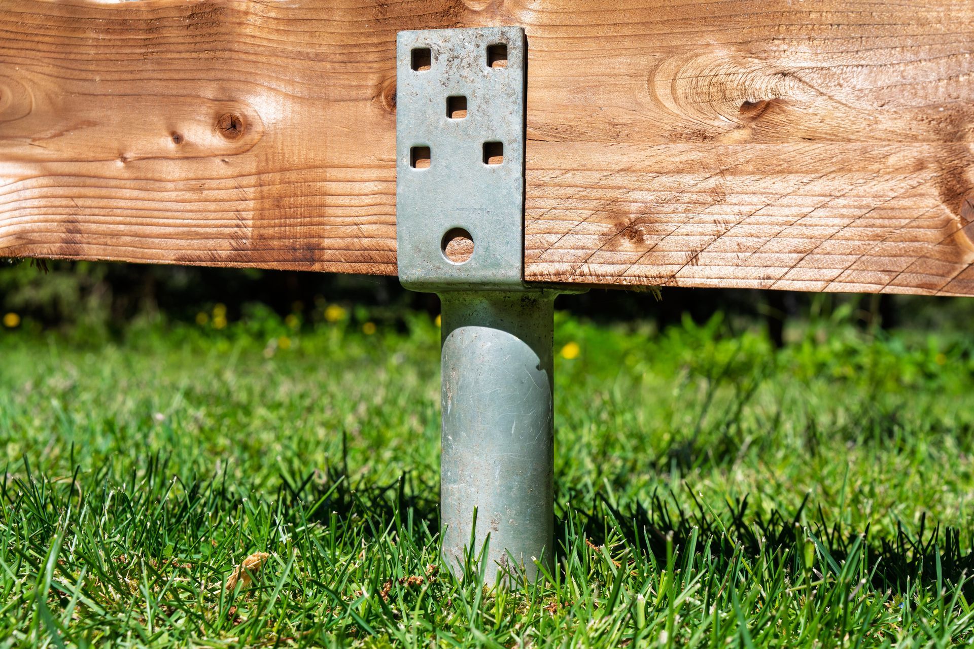 Metal post support under a wooden beam in grass, with a perforated galvanized bracket attached.