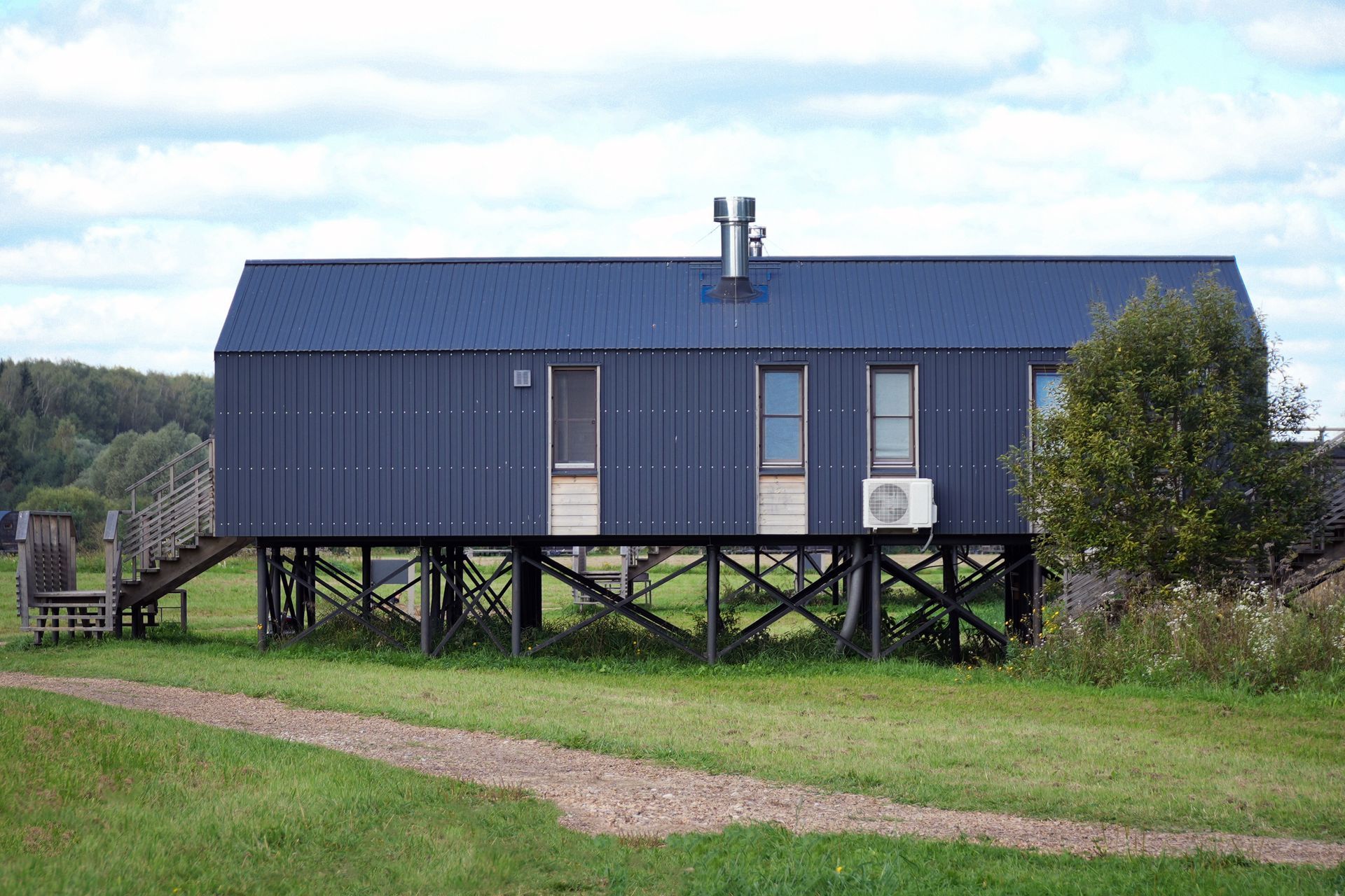 Blue house on stilts in a grassy field, with a gravel path and trees in the background