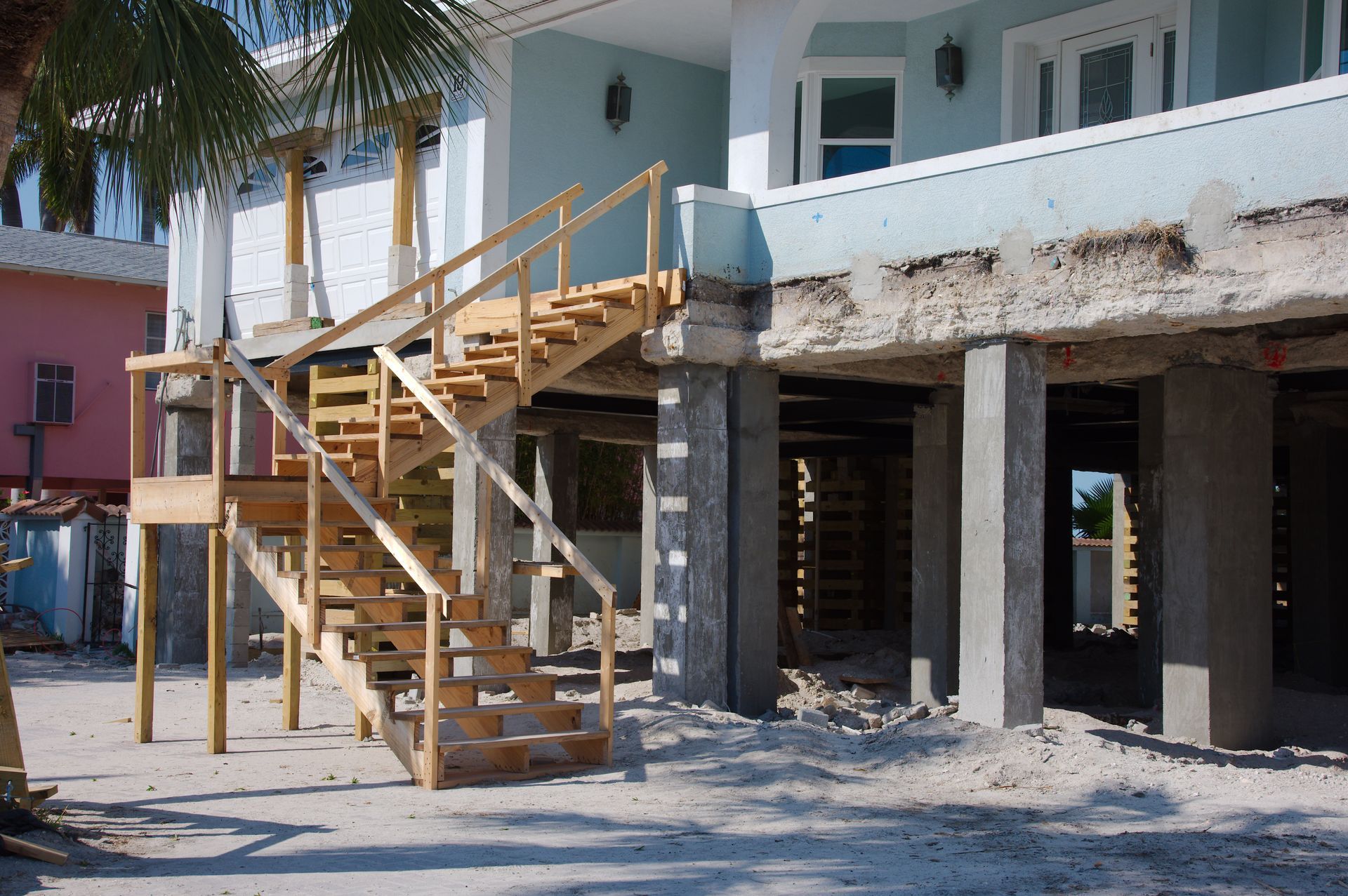 Wooden stairs leading to a raised beach house under construction beside palm trees and sand