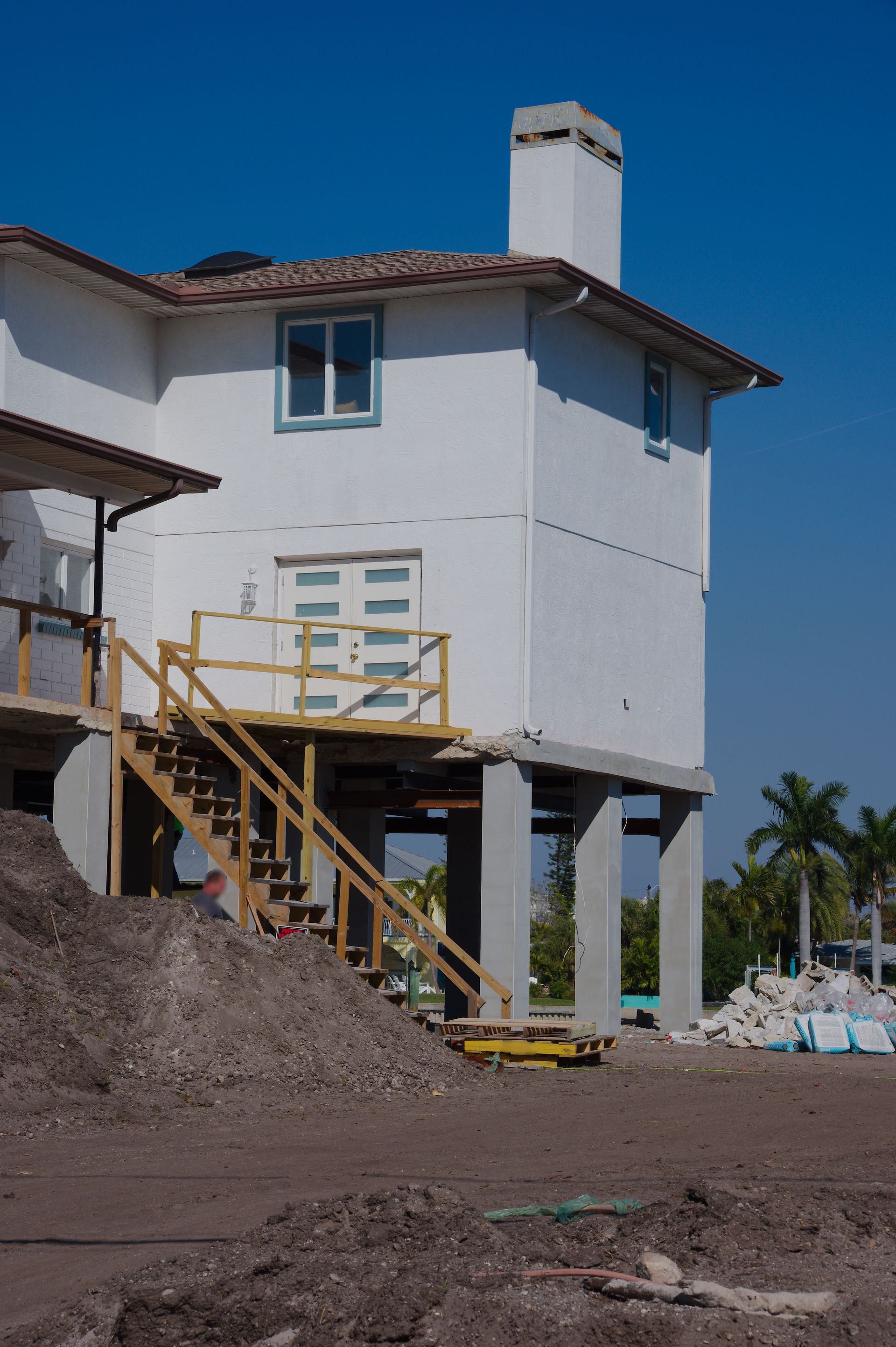 White two-story house under construction with stairs, porch railing, and a chimney on a dirt lot.