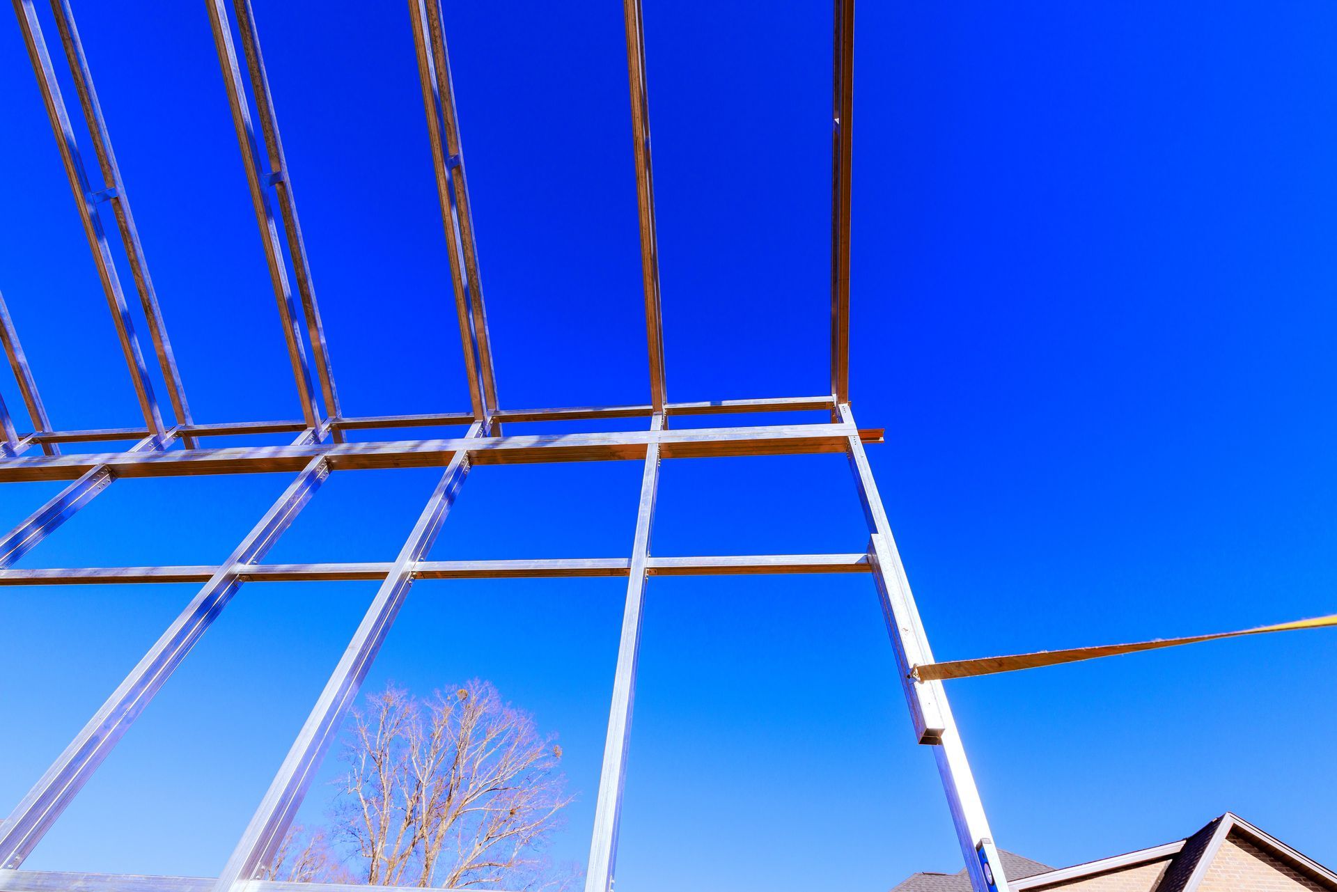Metal framing beams against a bright blue sky with a leafless tree and roofline below