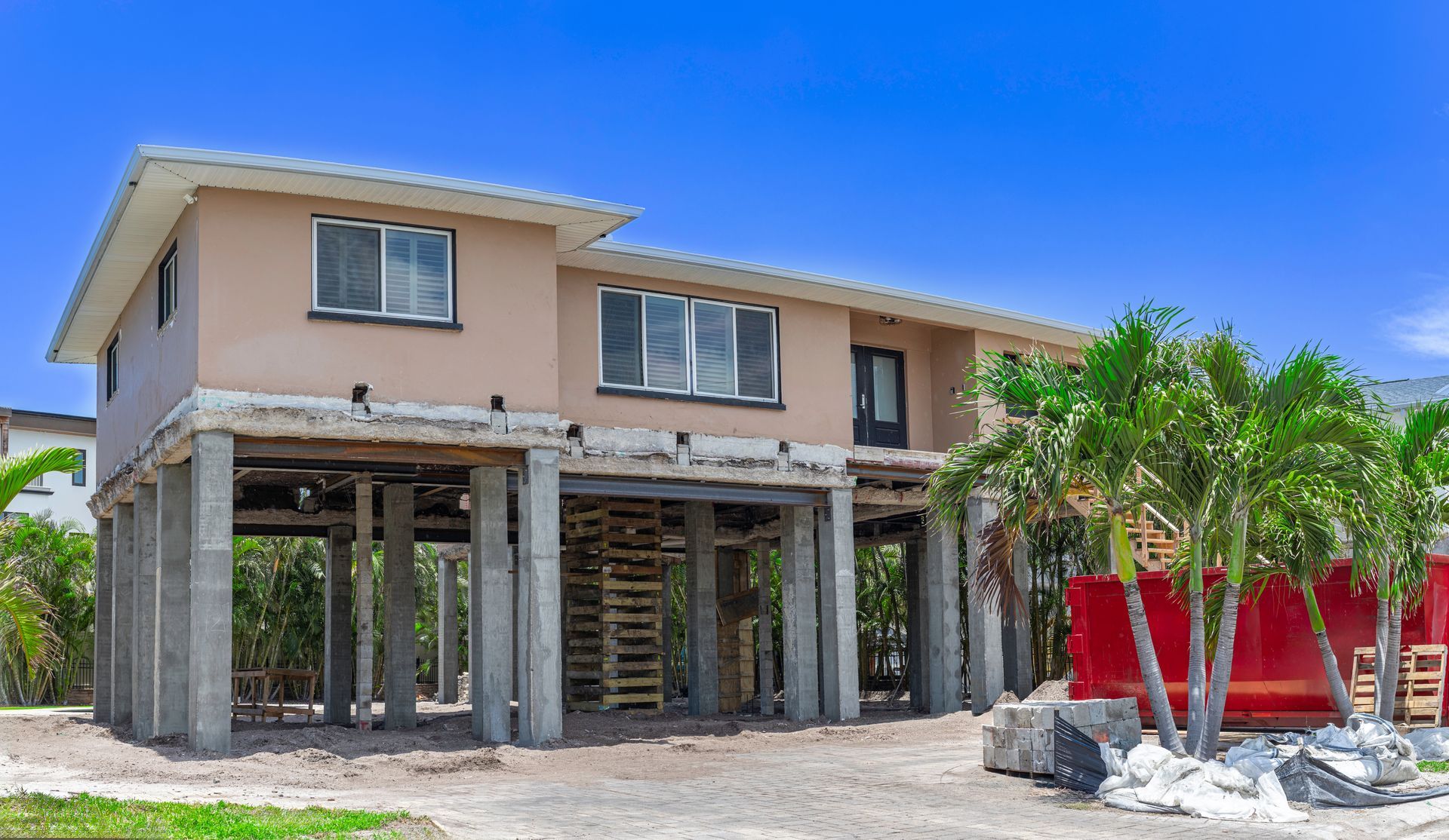 Two-story beige house with an unfinished ground floor, red fence, and palm trees under a blue sky