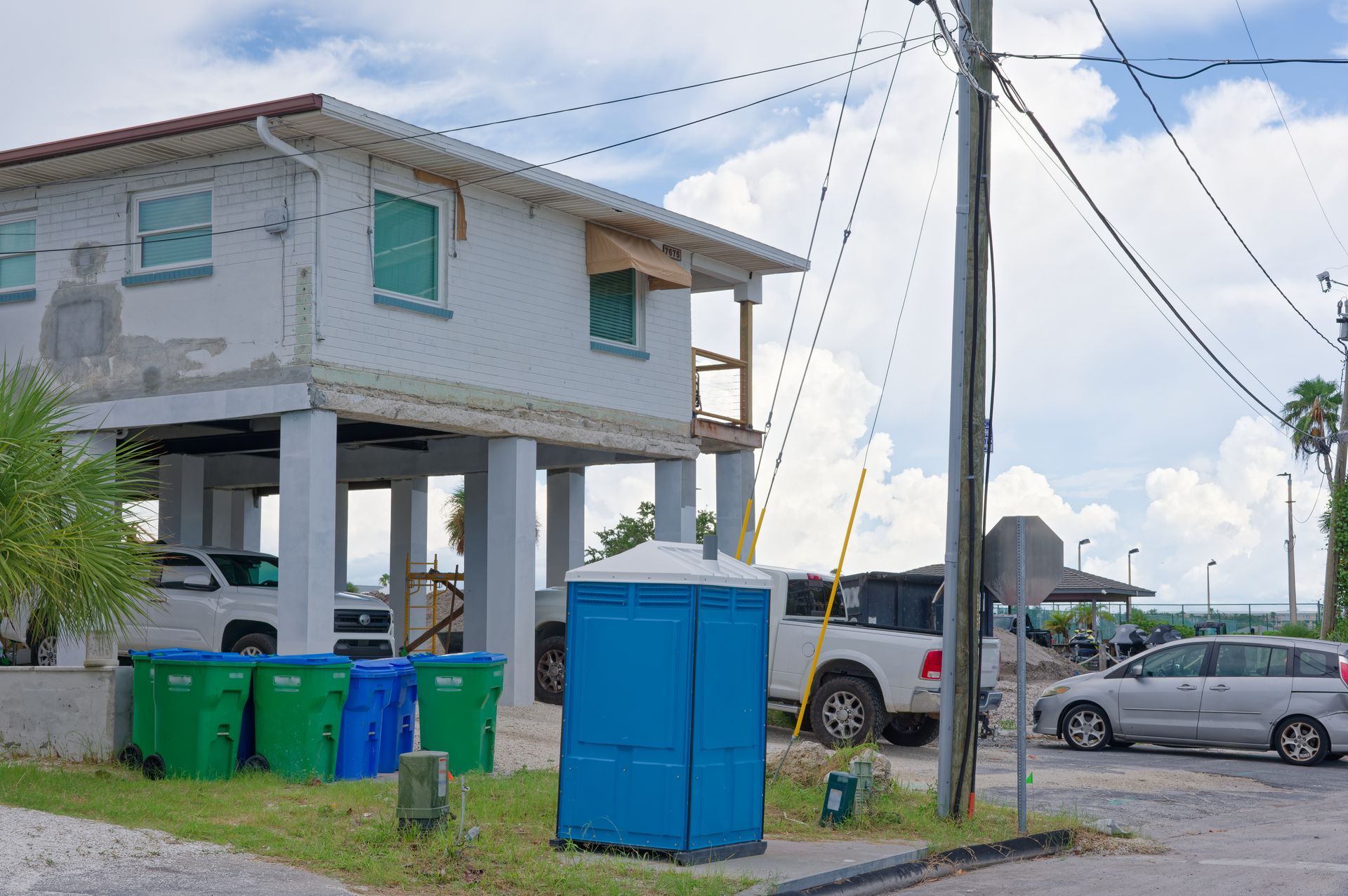 Raised house with parked cars and blue trash bins near a dirt lot